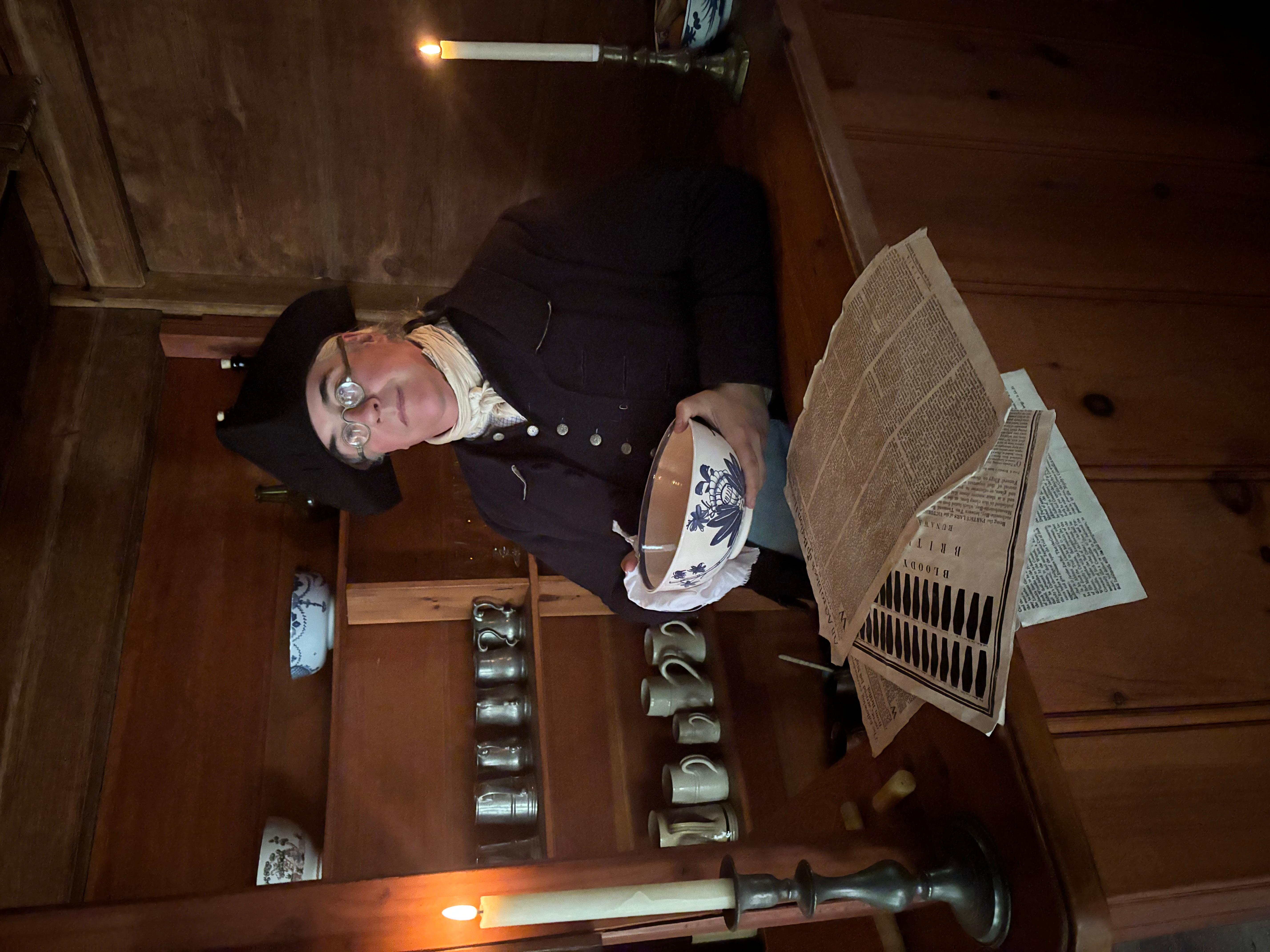 A man in colonial clothing stands behind a bar cleaning a punch bowl