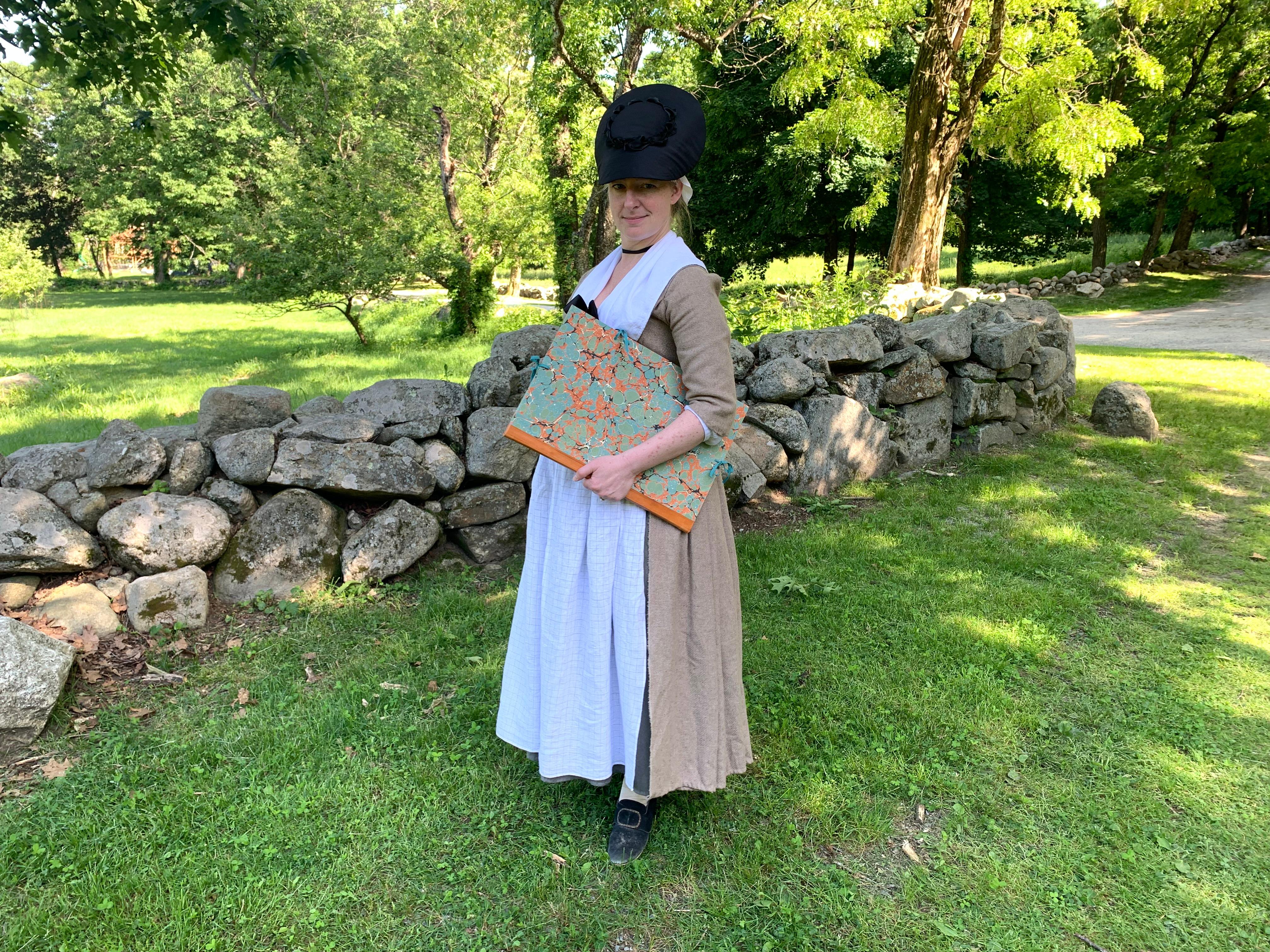 A woman in colonial clothing stands outside clutching a large book.