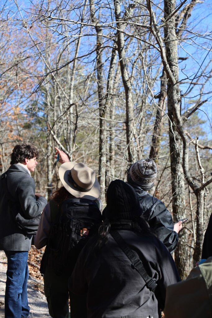 A park ranger in a grey hat points into the trees alongside a group of hikers.