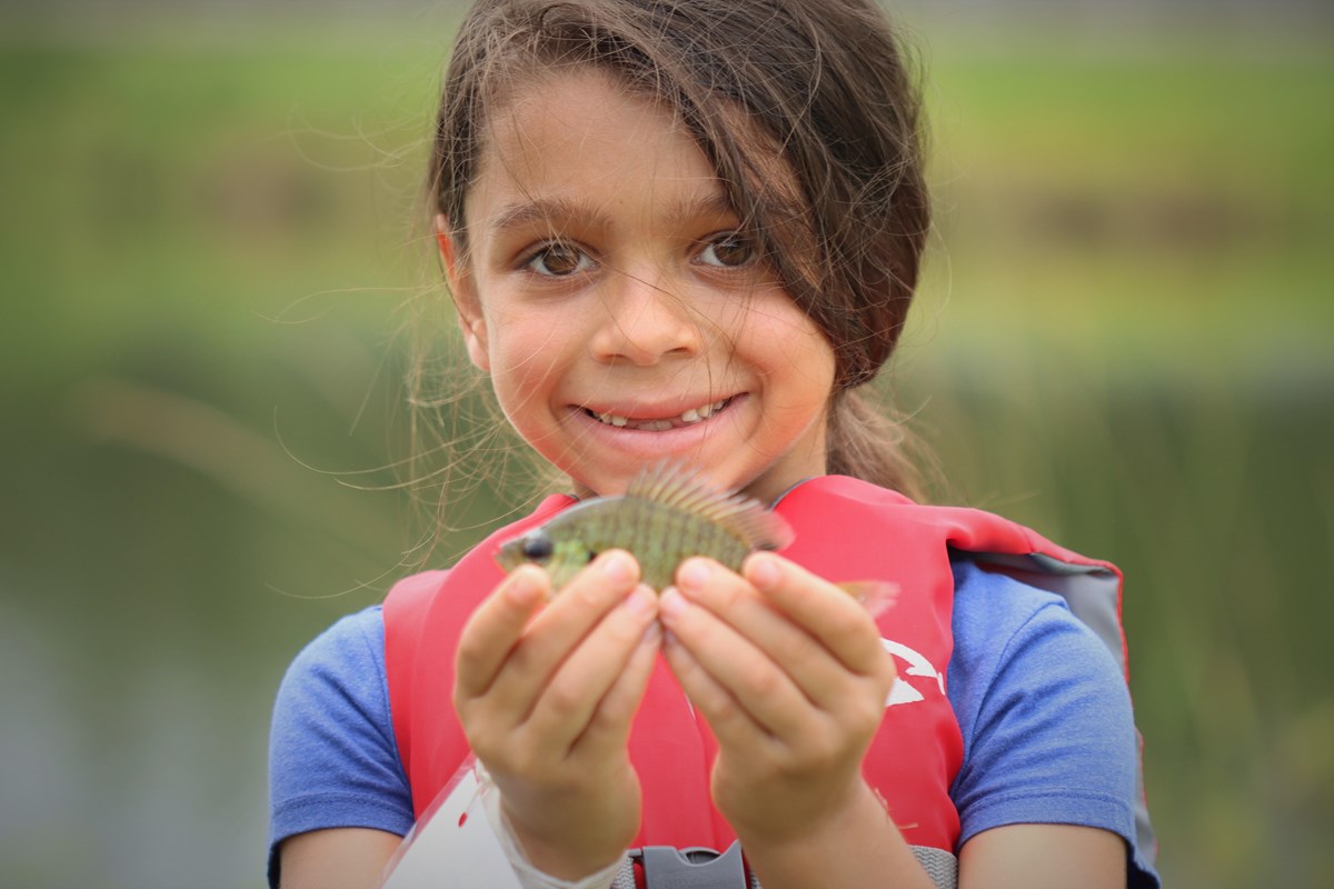 young girl smiling while holding up a small green fish that she caught.