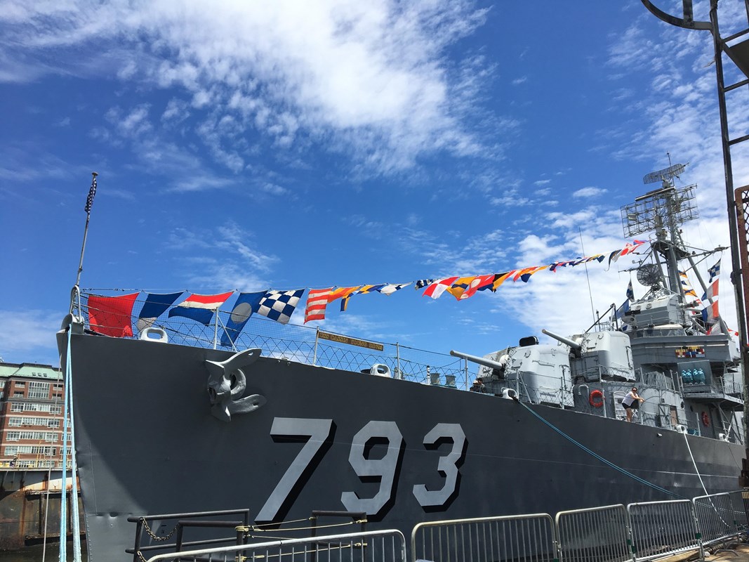 The bow of the USS CASSIN YOUNG with flags hangs from a rope connected to the top of the ship.