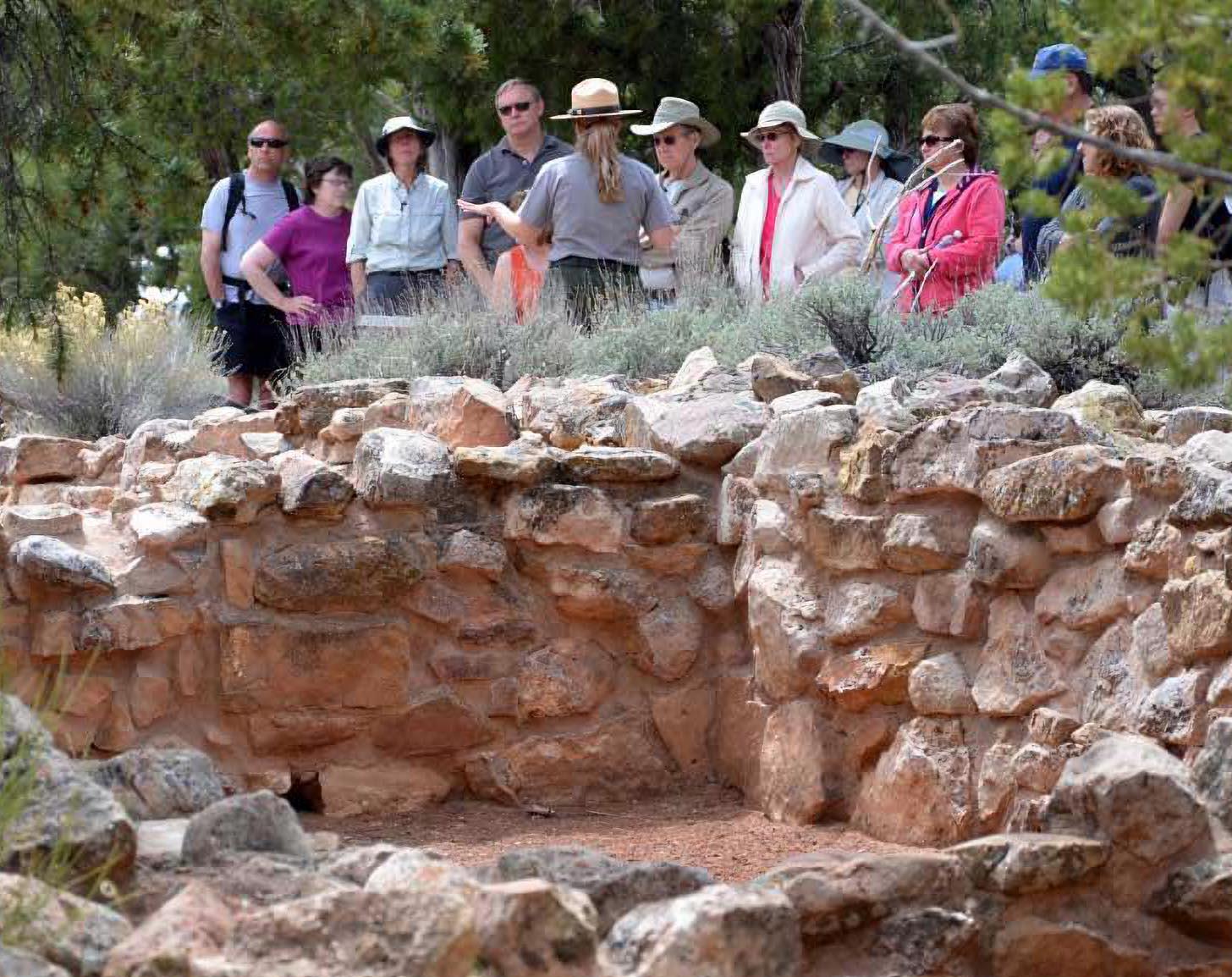 A park ranger is giving a talk to a dozen people with a partial stone wall of an archaeology site.