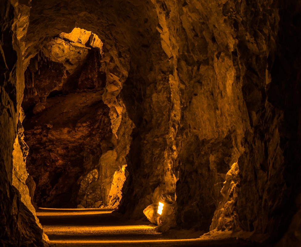 A dimly lit, underground tunnel with rocky walls and a flat floor leads to further passages