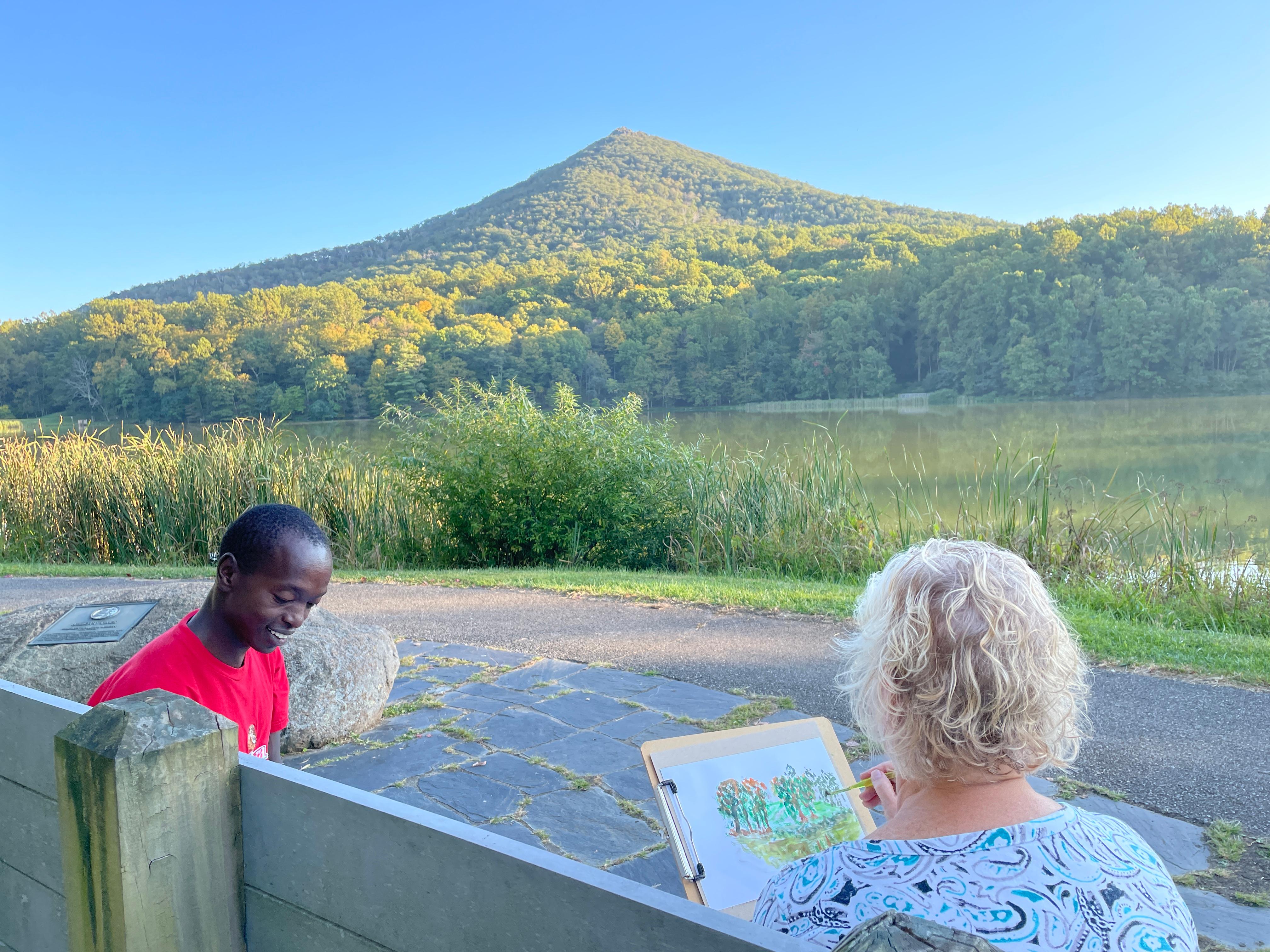 a man & woman sit on a bench painting with a lake and mountain in the background