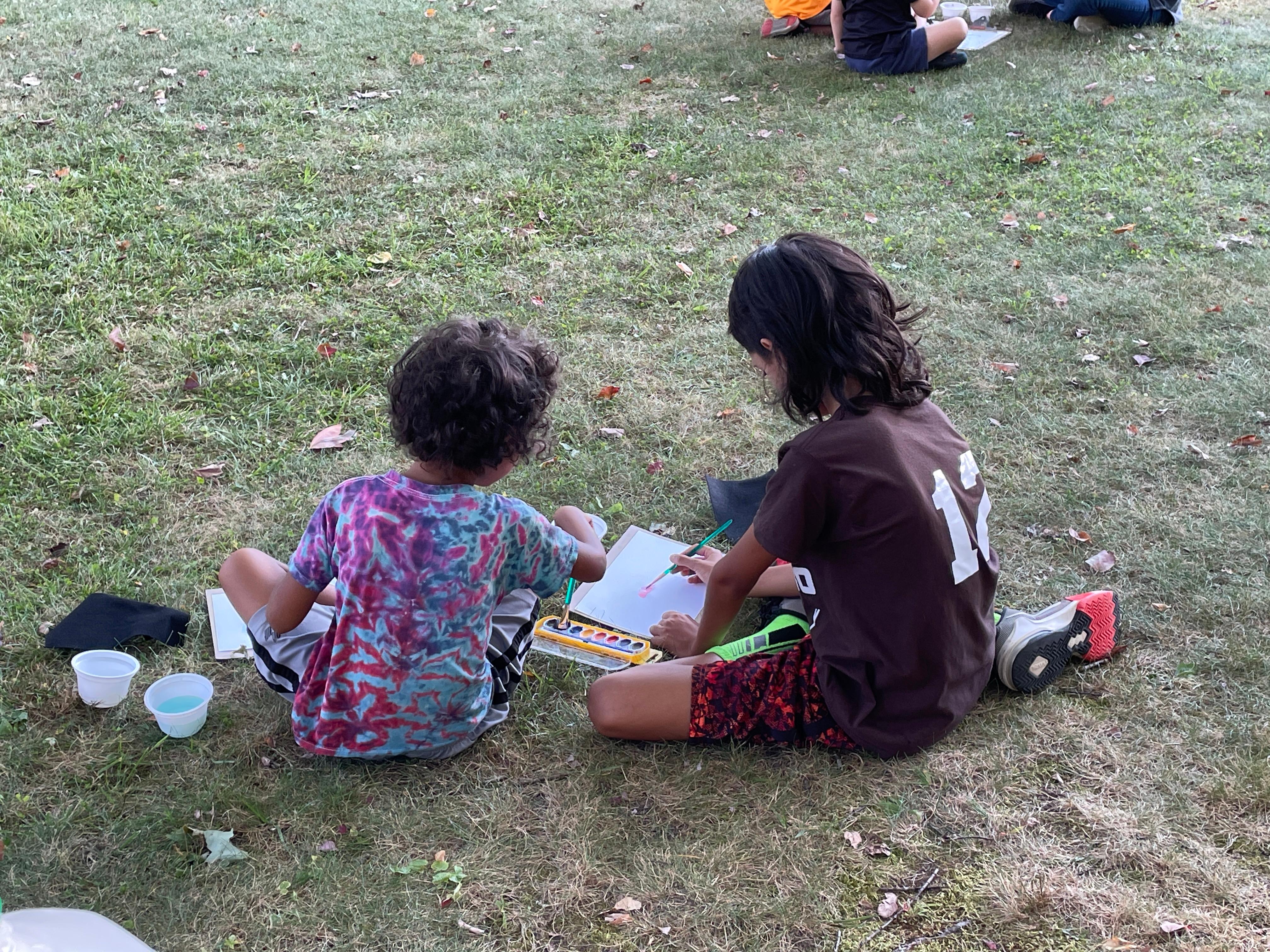 backs of 2 kids sitting in the grass painting with watercolors