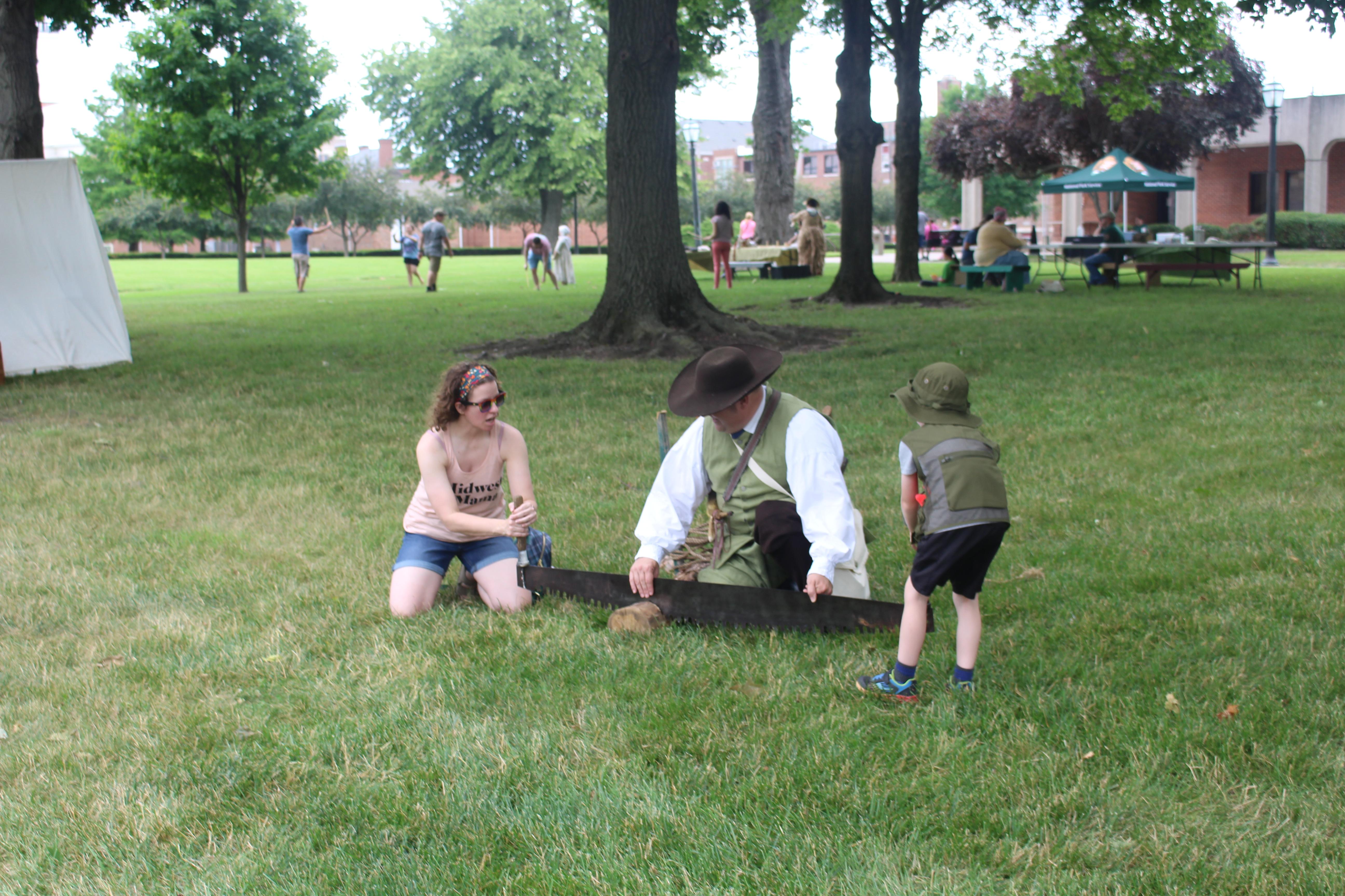An adult and child saw a log with the help of a ranger dressed in 18th century style clothing