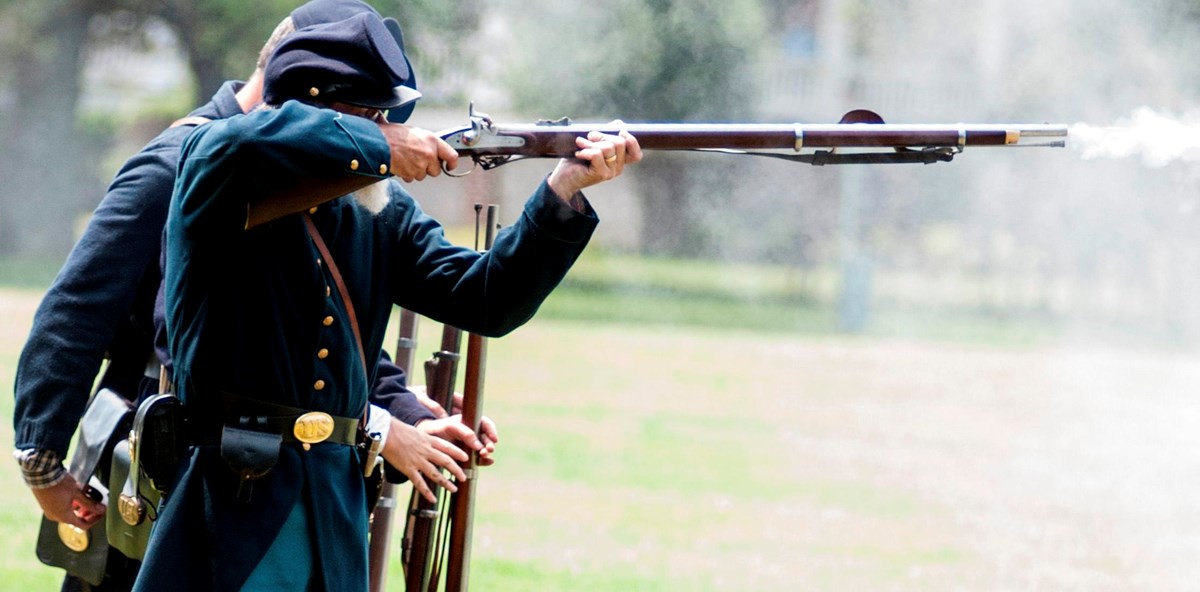 US Soldiers in American Civil War uniforms stand in a line firing muskets.
