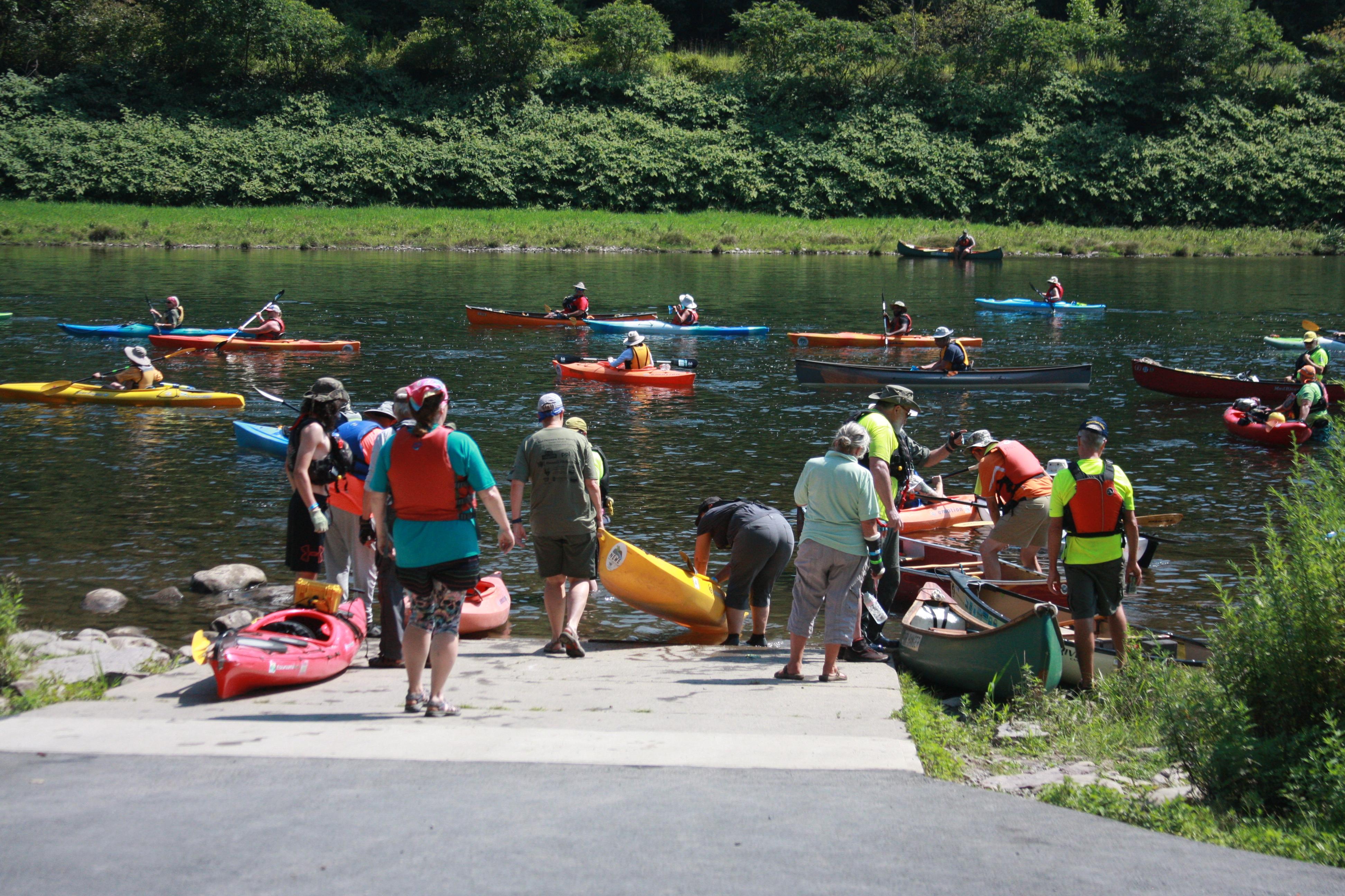 People carry and take off colorful canoes and kayaks at a concrete boat ramp into the river.