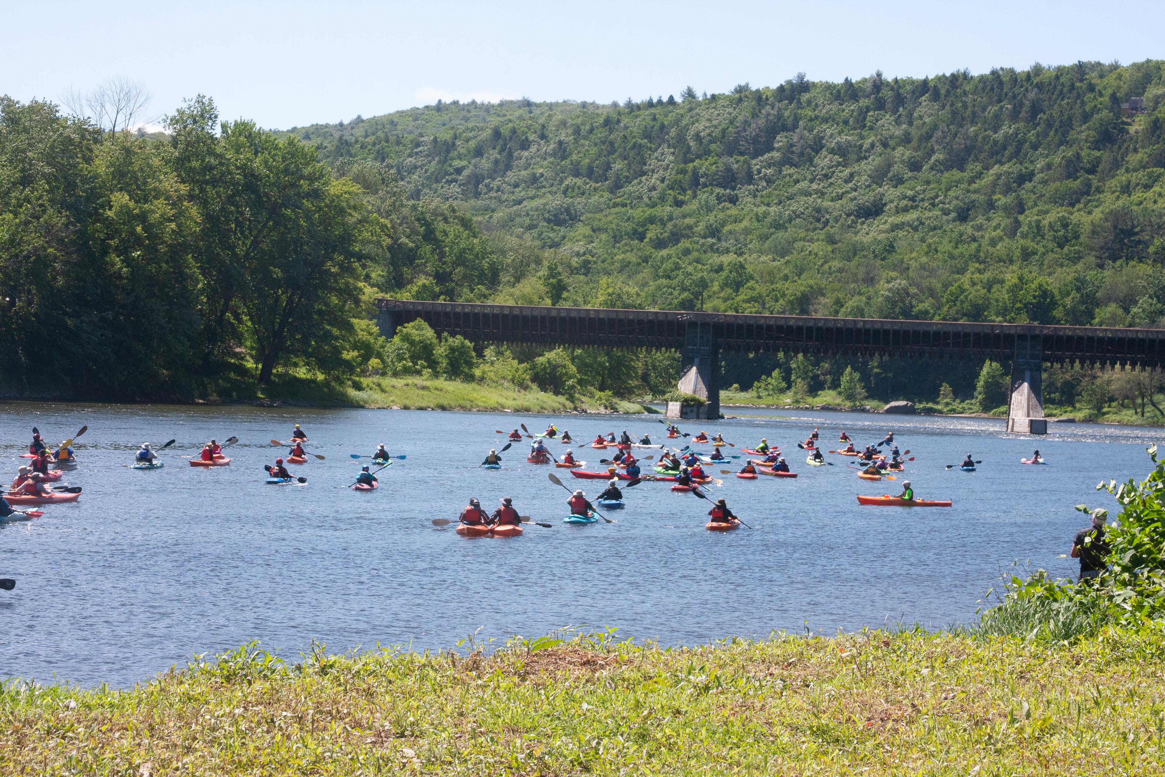Approximately 50 people in individual kayaks and canoes float on the river. Bridge in distance.