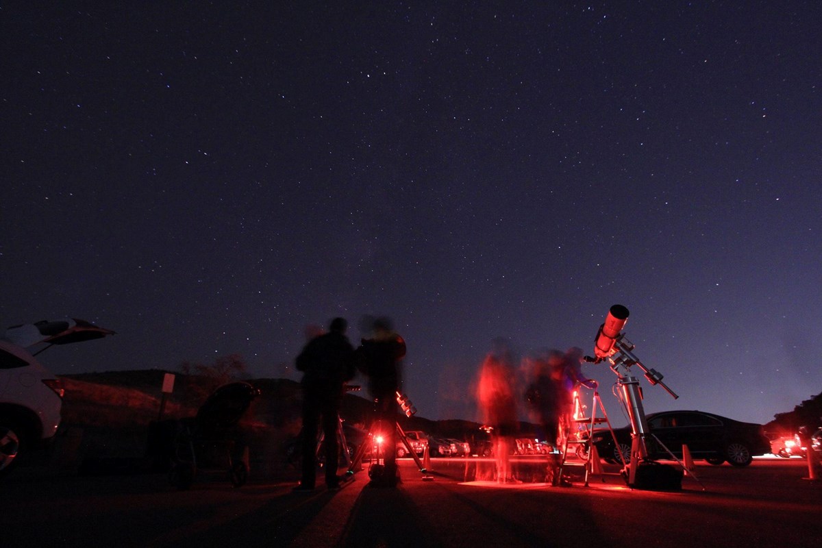 Starry night sky with telescope and people moving in red light in the foreground.