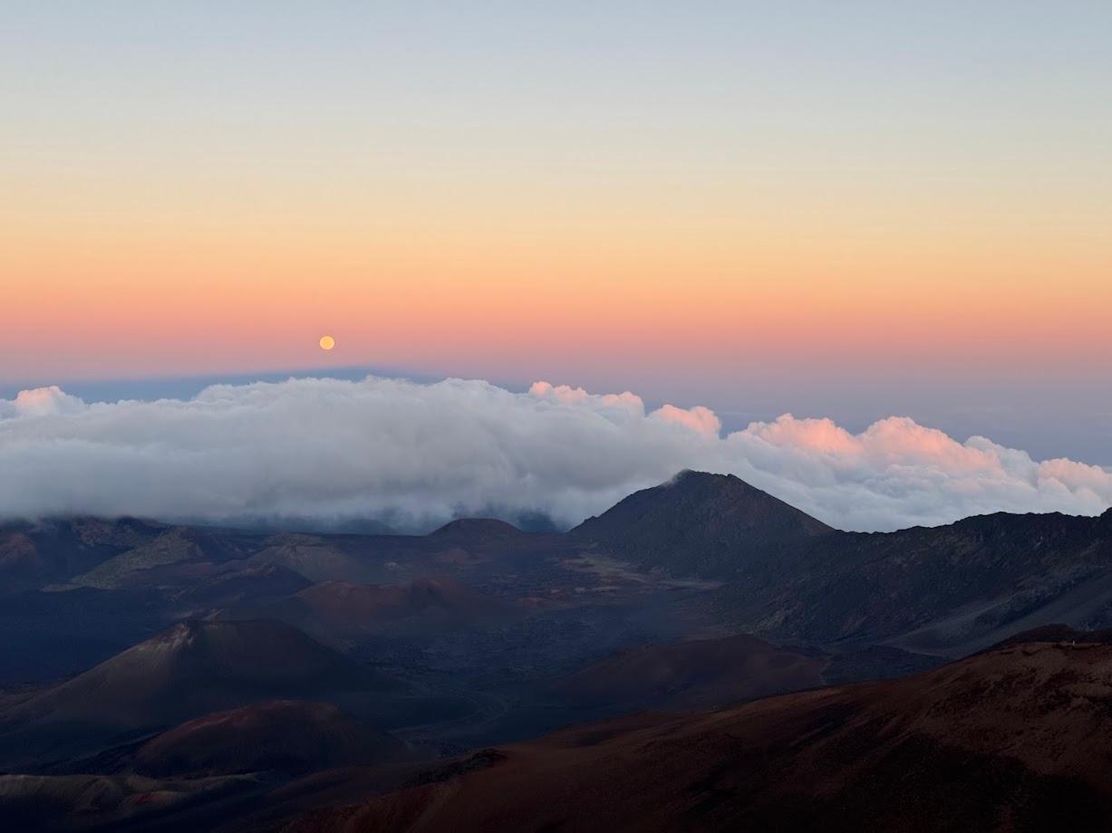 The moon rises amid a pink sky, with clouds and a volcanic landscape below