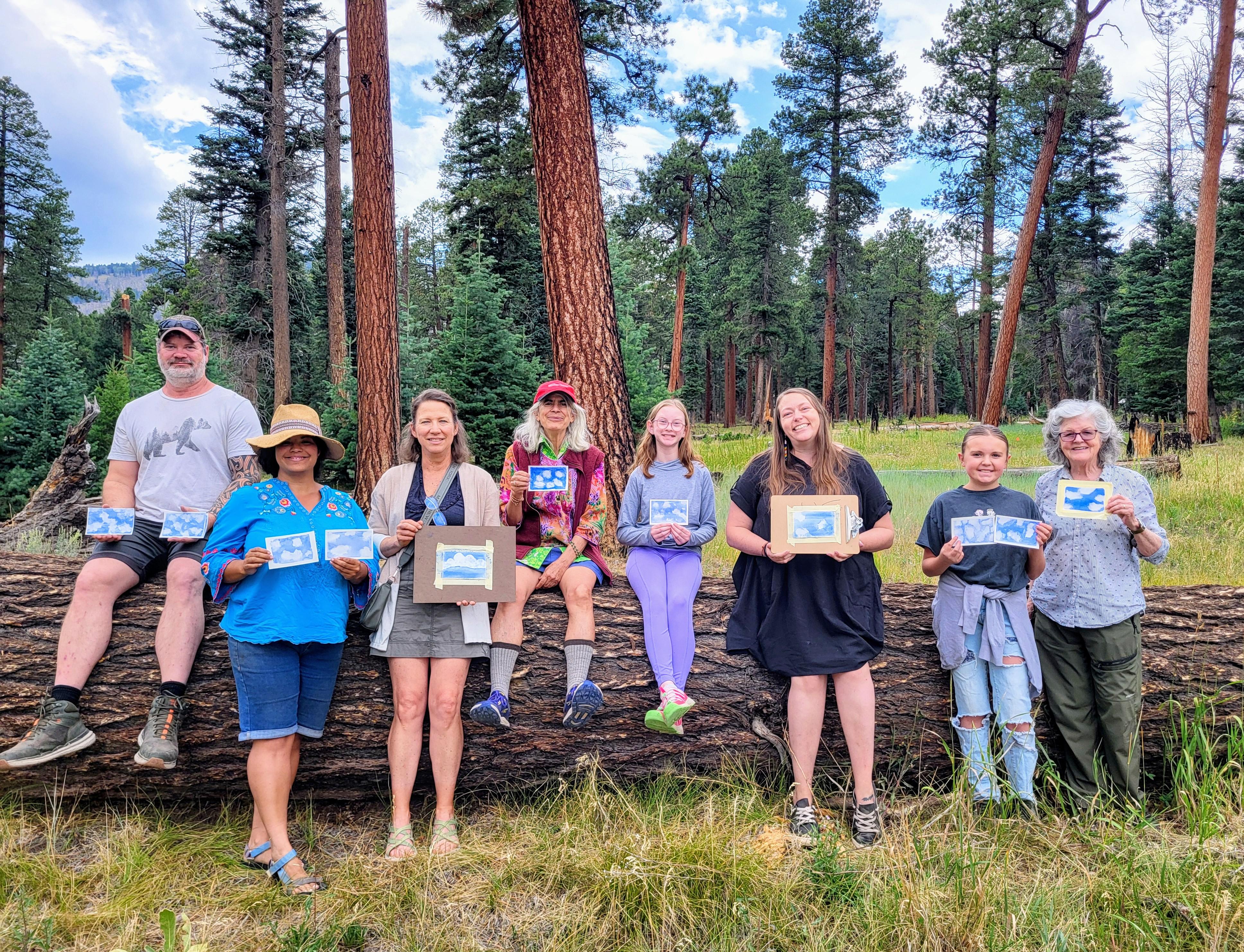 A group of artists stand outdoors with small paintings of clouds in the sky.
