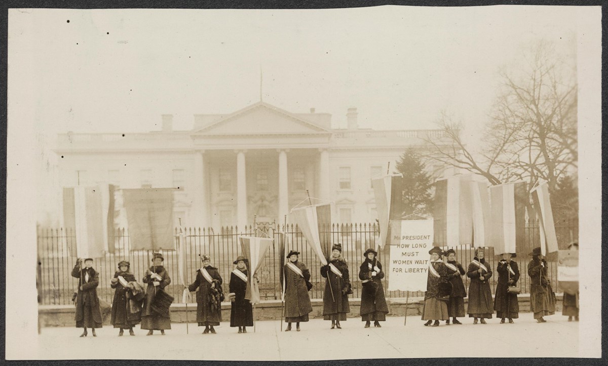 Row of about 15 women wearing suffrage sashes and holding banners standing in front of White House