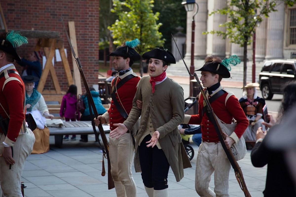 Color photo of a man in colonial costume being led away by British soldiers in red military uniforms