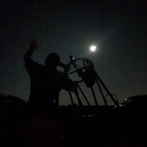 Night photograph silhouetting someone looking through a telescope at the bright white moon.
