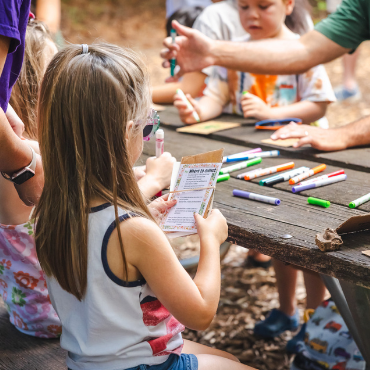 Children and an adult gathered around a picnic table with markers and crafts.