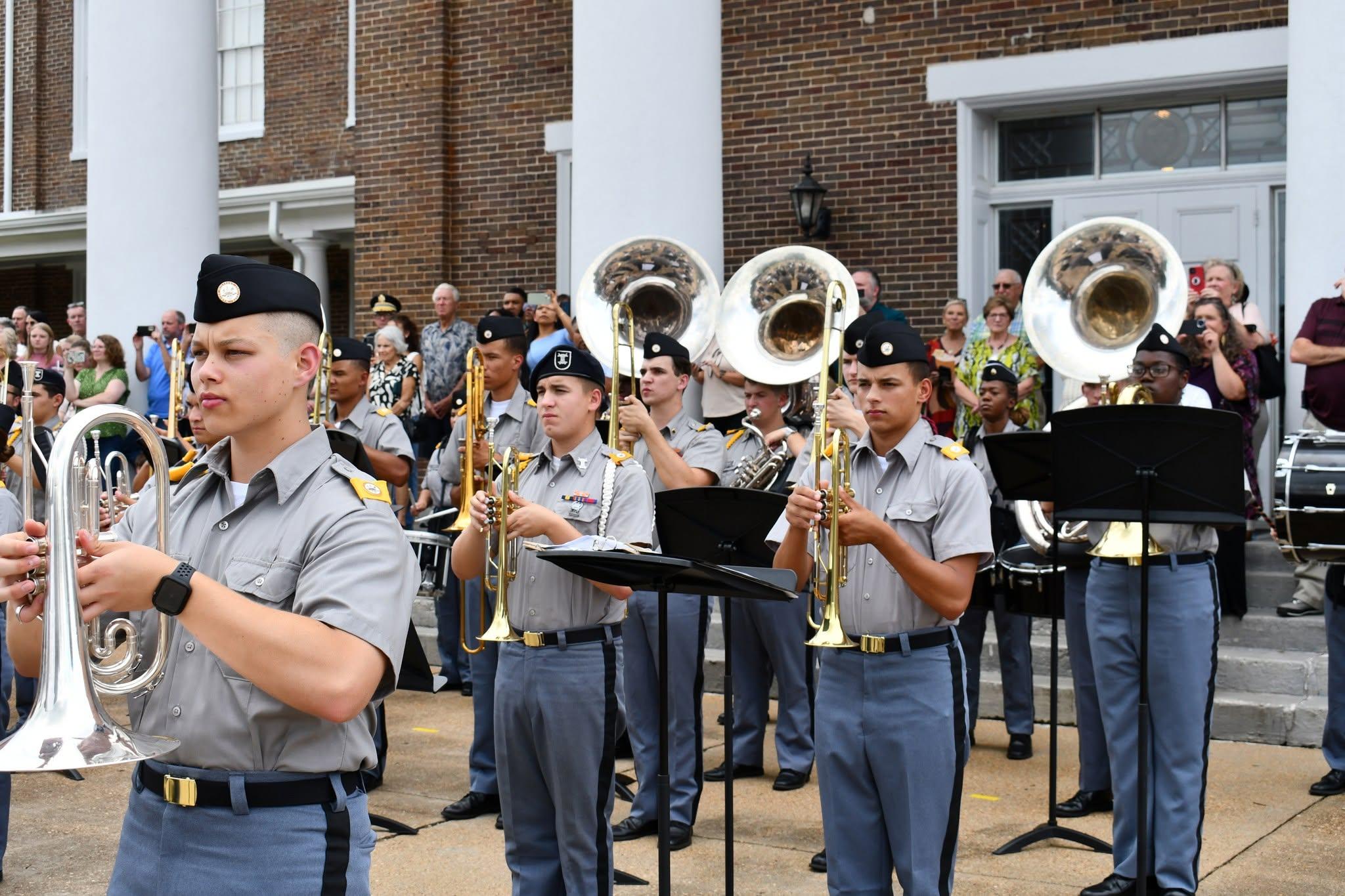 Military cadet band holding instruments in front of a red brick building with white columns