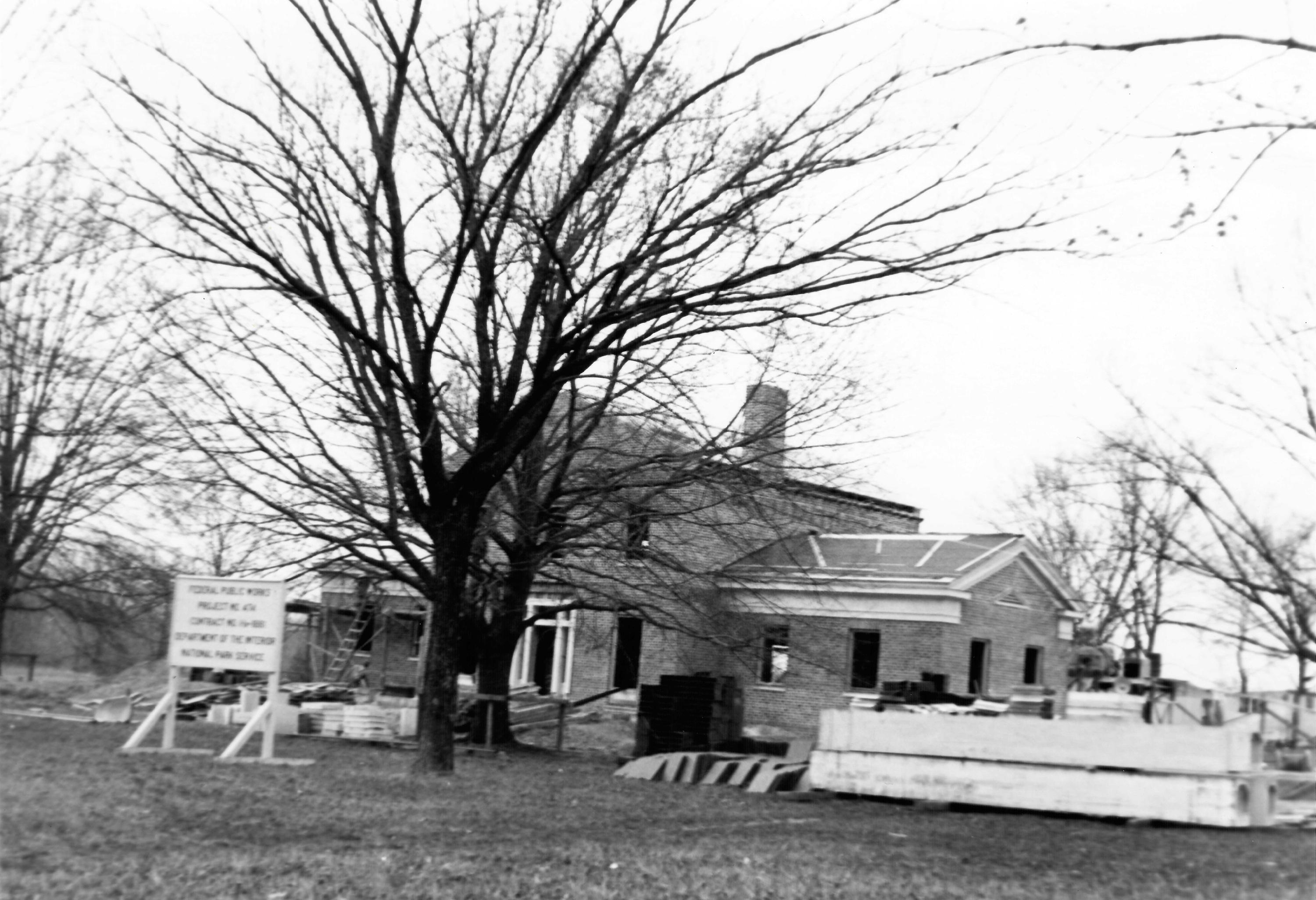 A black‑and‑white photograph of a brick building under construction, surrounded by leafless trees.