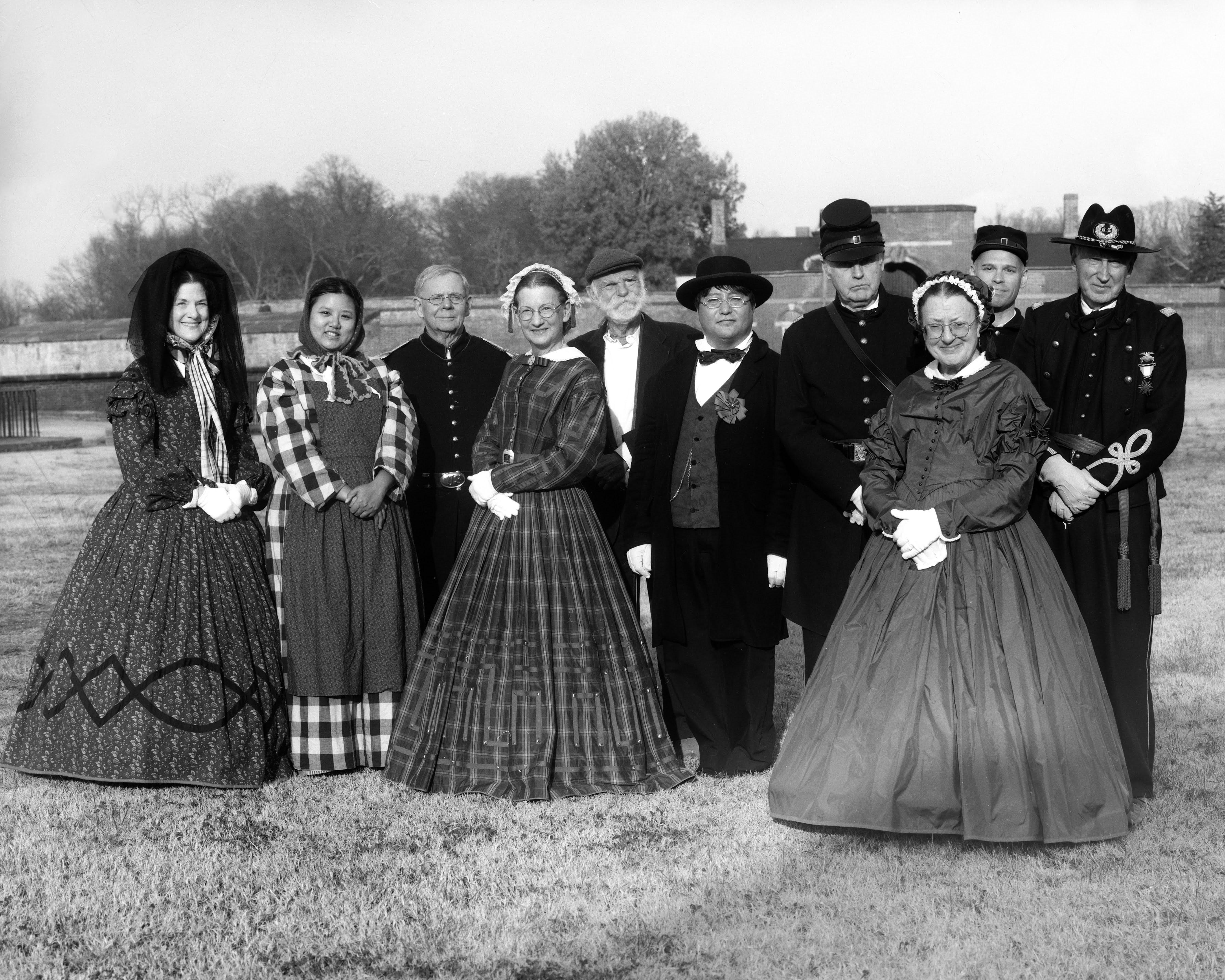 A black white image of dancers in period accurate dresses and uniforms standing shoulder to shoulder