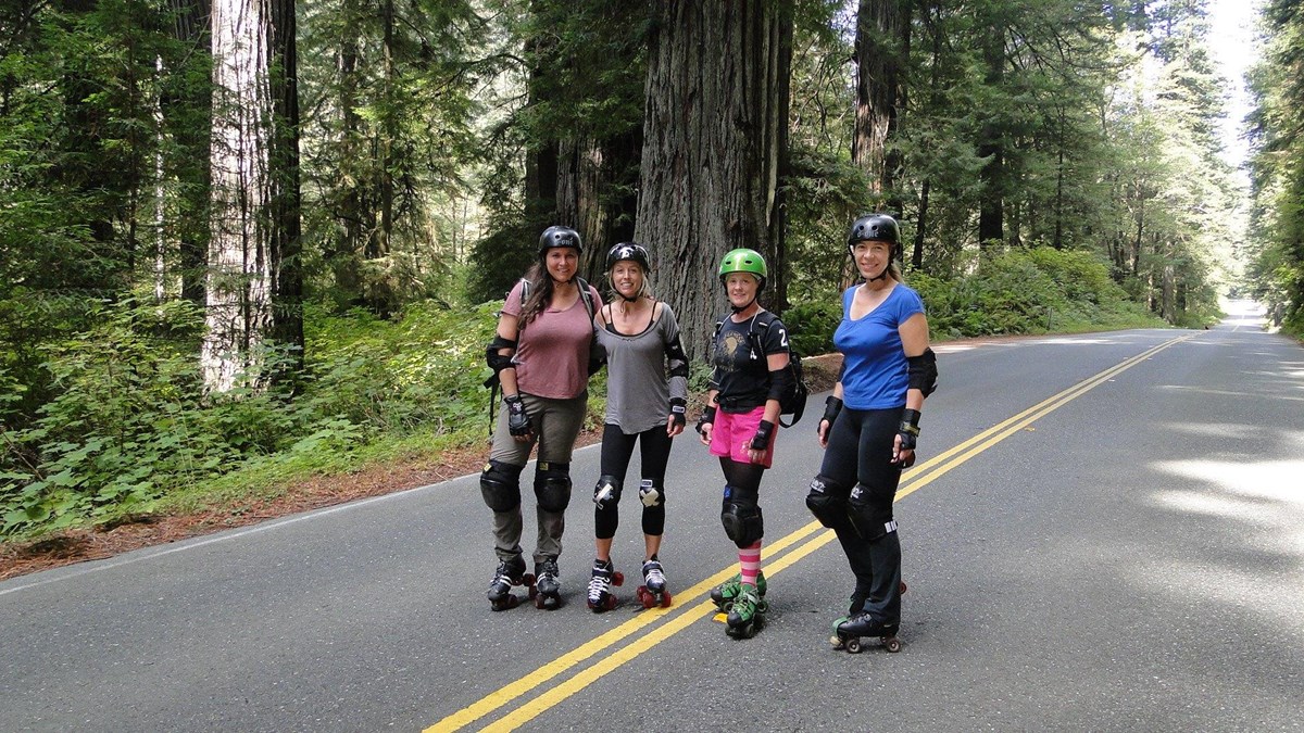 Four women in skating clothes stand in the middle of a road lined with redwood trees.