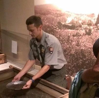 A man in a ranger uniform holds a silver pan in a wooden box demonstrating how to gold pan.