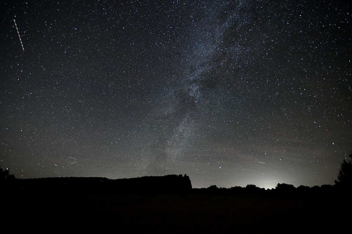The Milky Way extends across the sky and behind a silhouetted mesa.