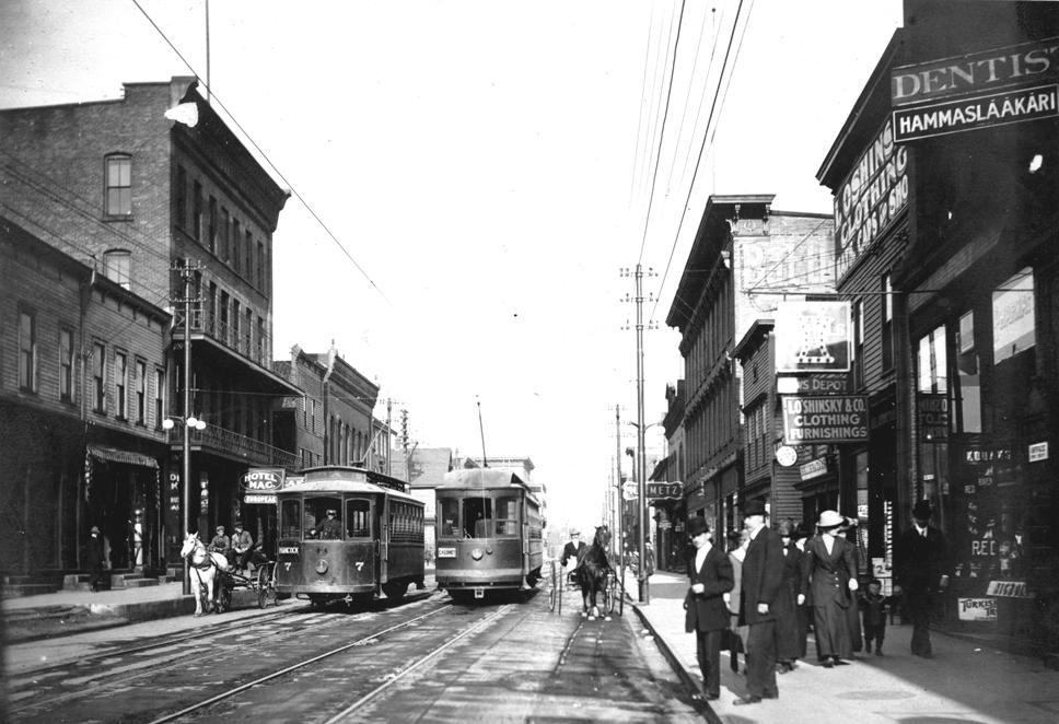 Early 20th-century city street with two streetcars, pedestrians, horse and carriage and storefronts.