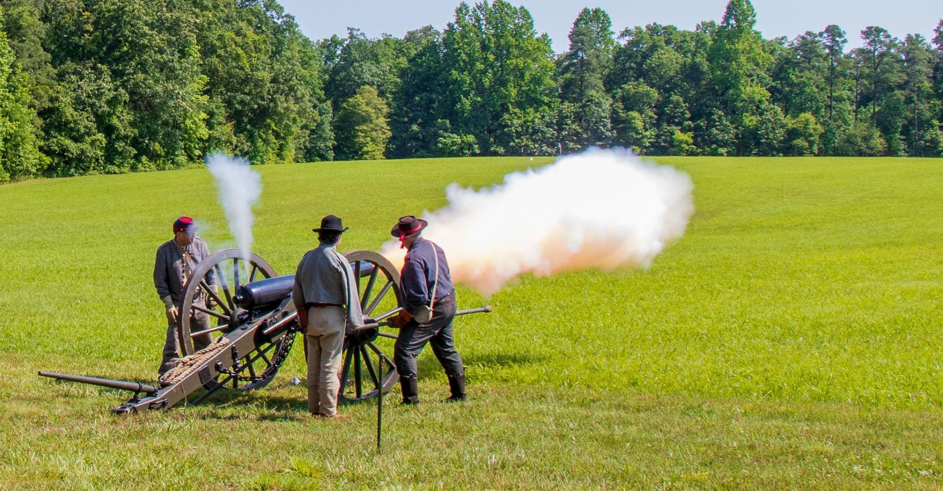 Three men in gray wool Civil War uniforms firing a black, iron cannon