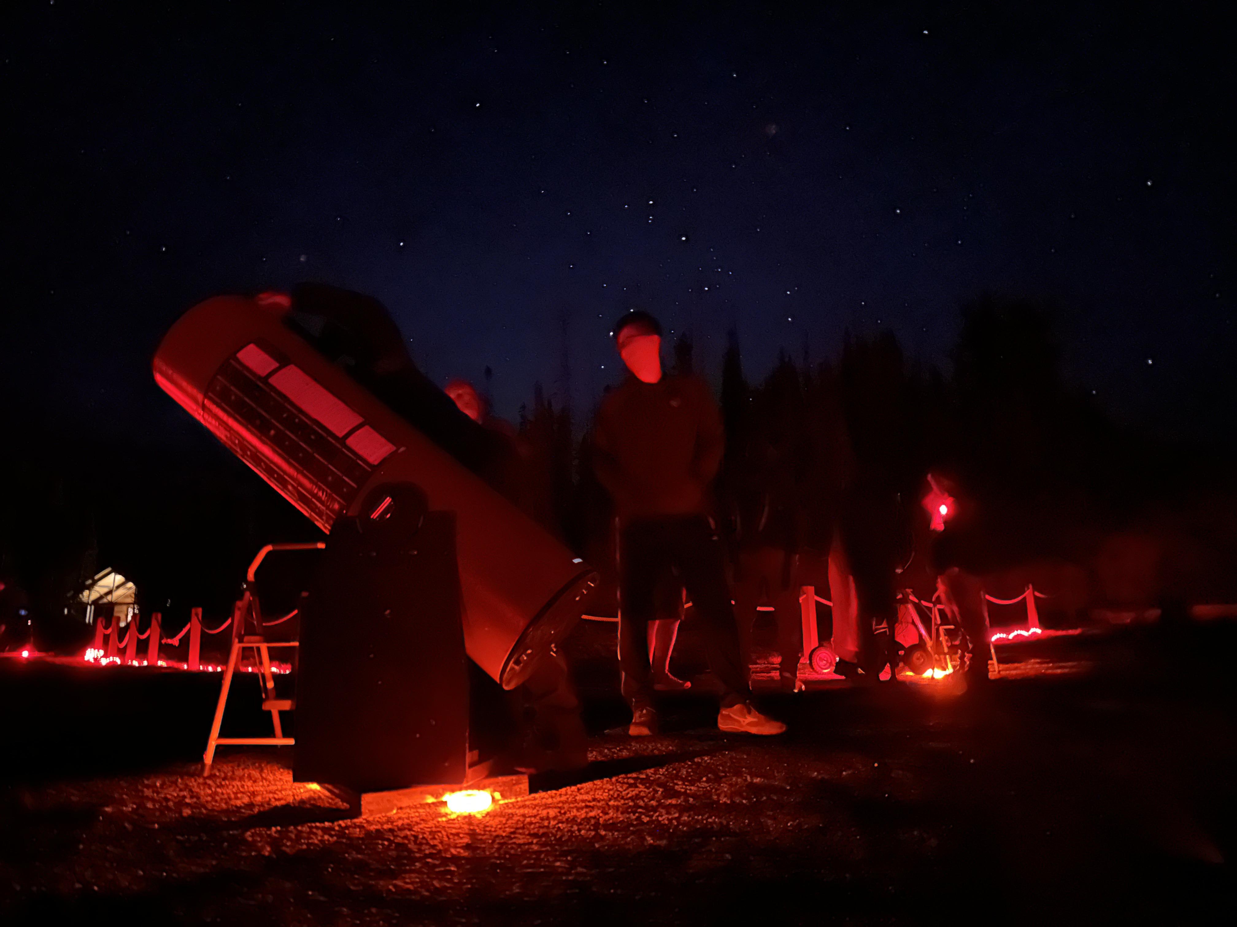 A visitor stands next to a large telescope in the warm glow of red light with a starry backdrop.