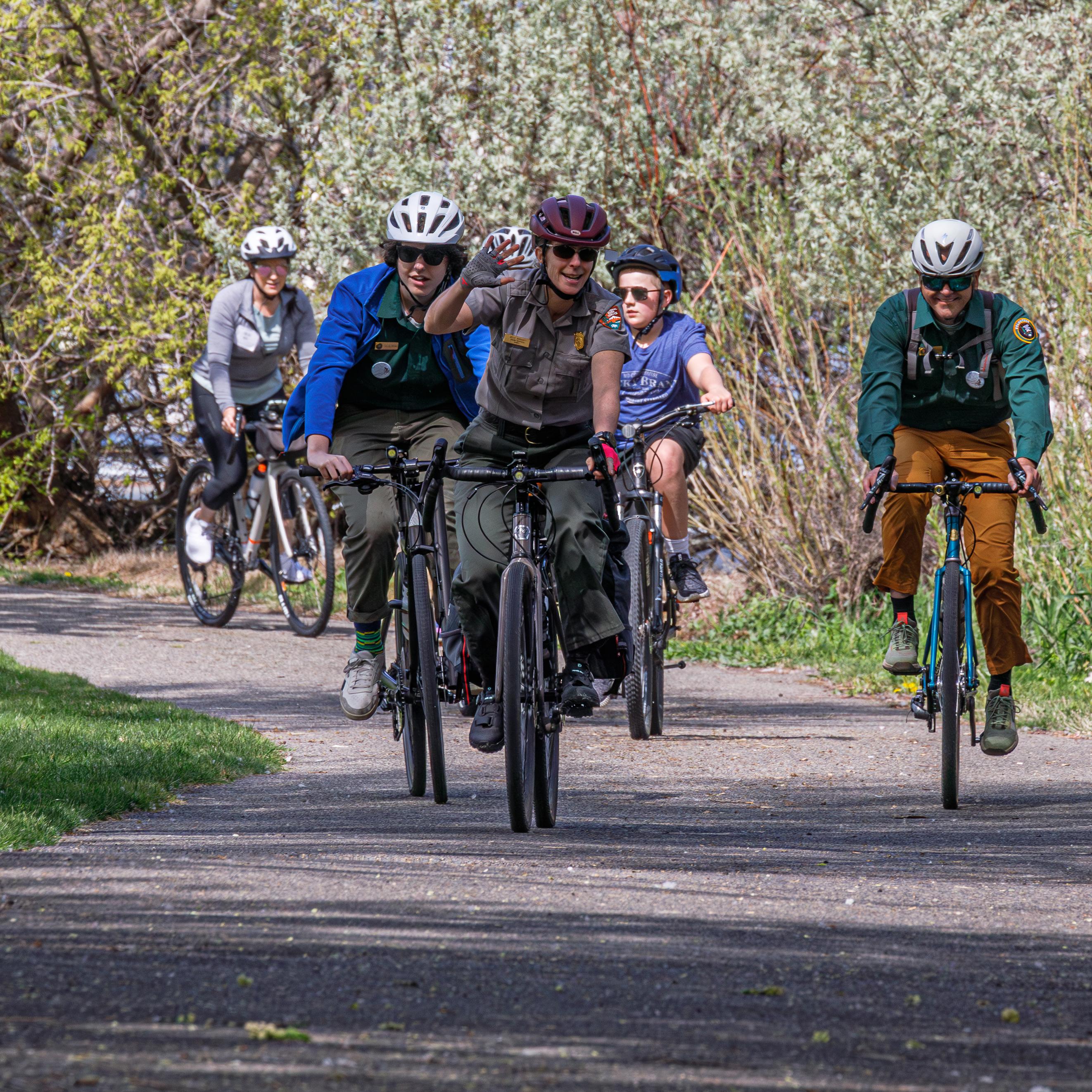 5 cyclists ride along a paved path in a park with grass and trees.