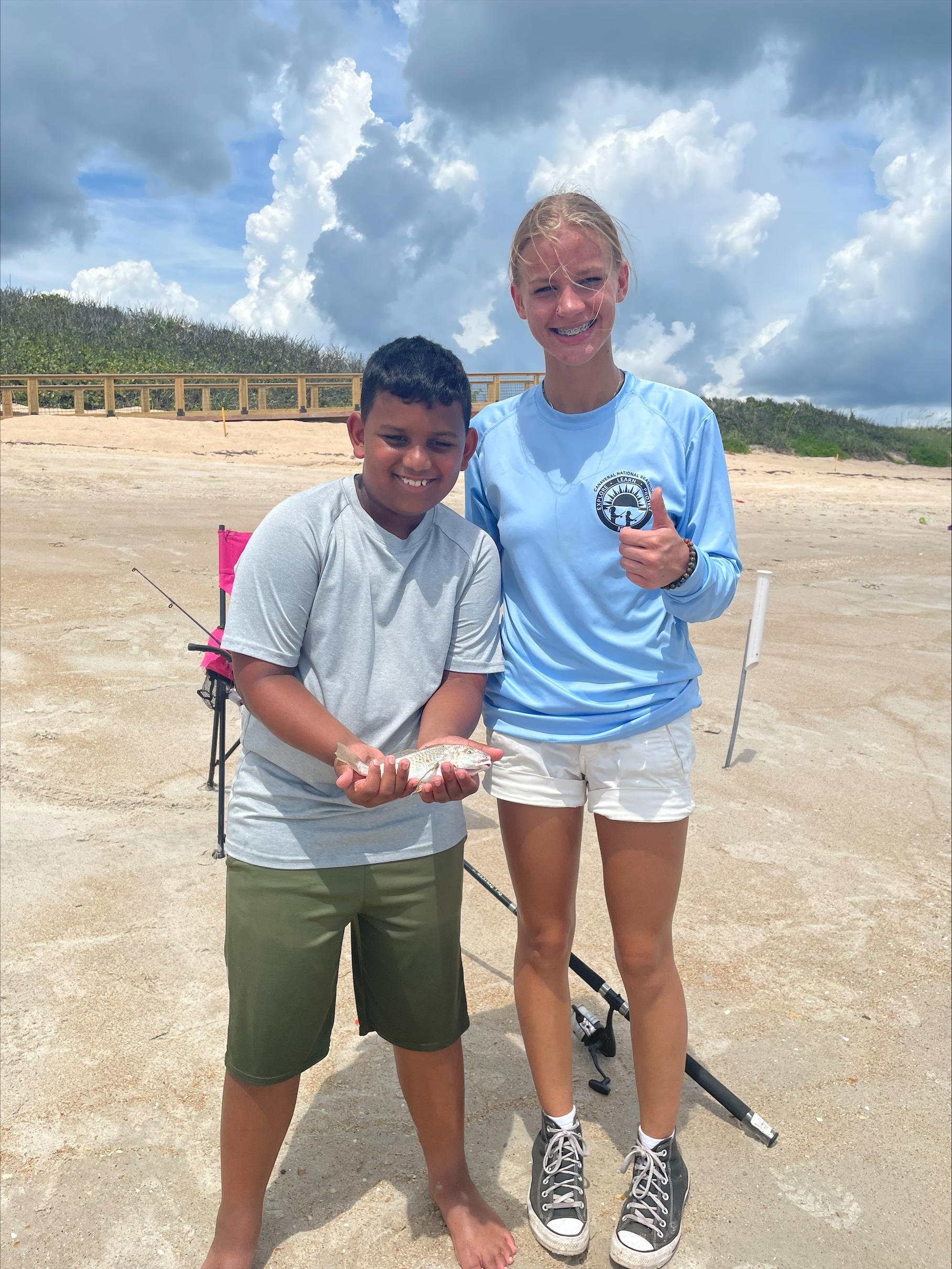 A Jr Angler is holding a fish standing next to an instructor on the beach.