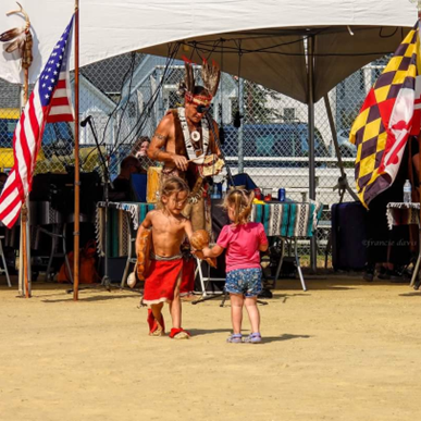 Children at a cultural event with a man in traditional attire under a canopy with flags.