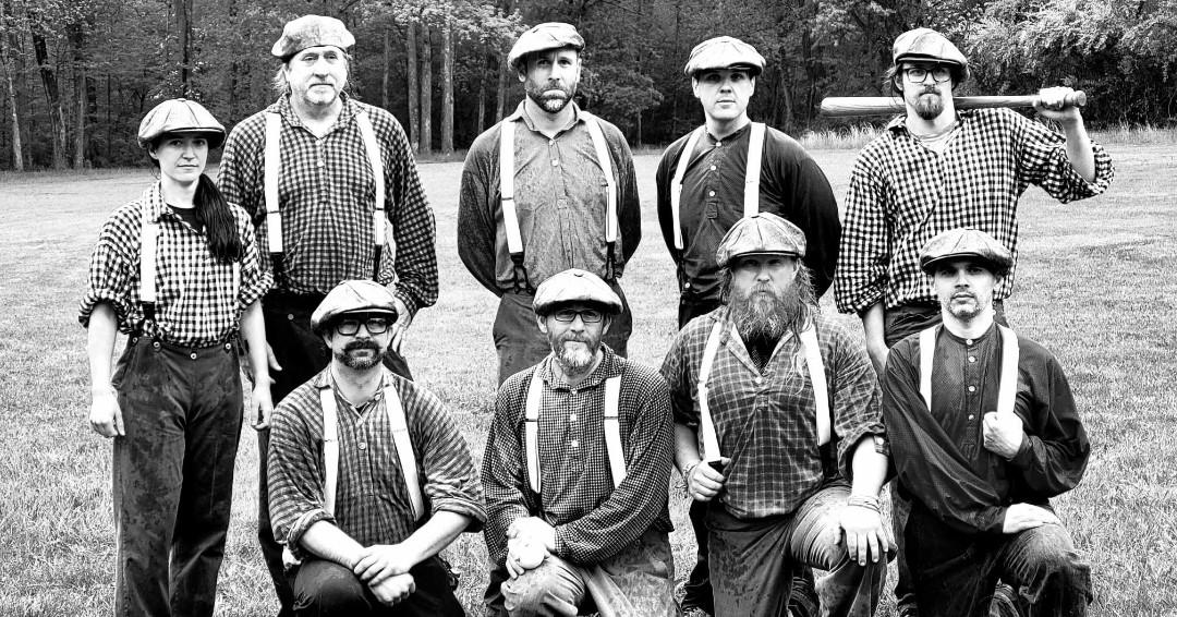 Black and white photo of people in historic attire, one holding a baseball bat.