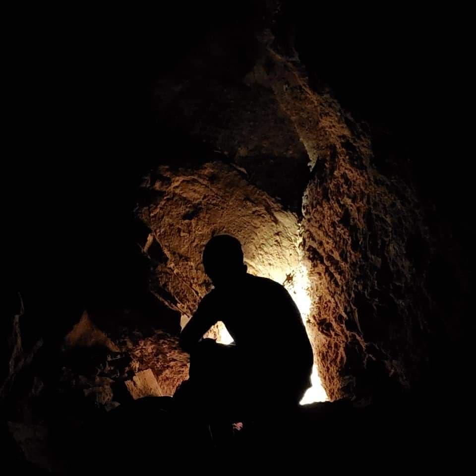 The silhouette of a person wearing caving equipment near a small lit passageway.