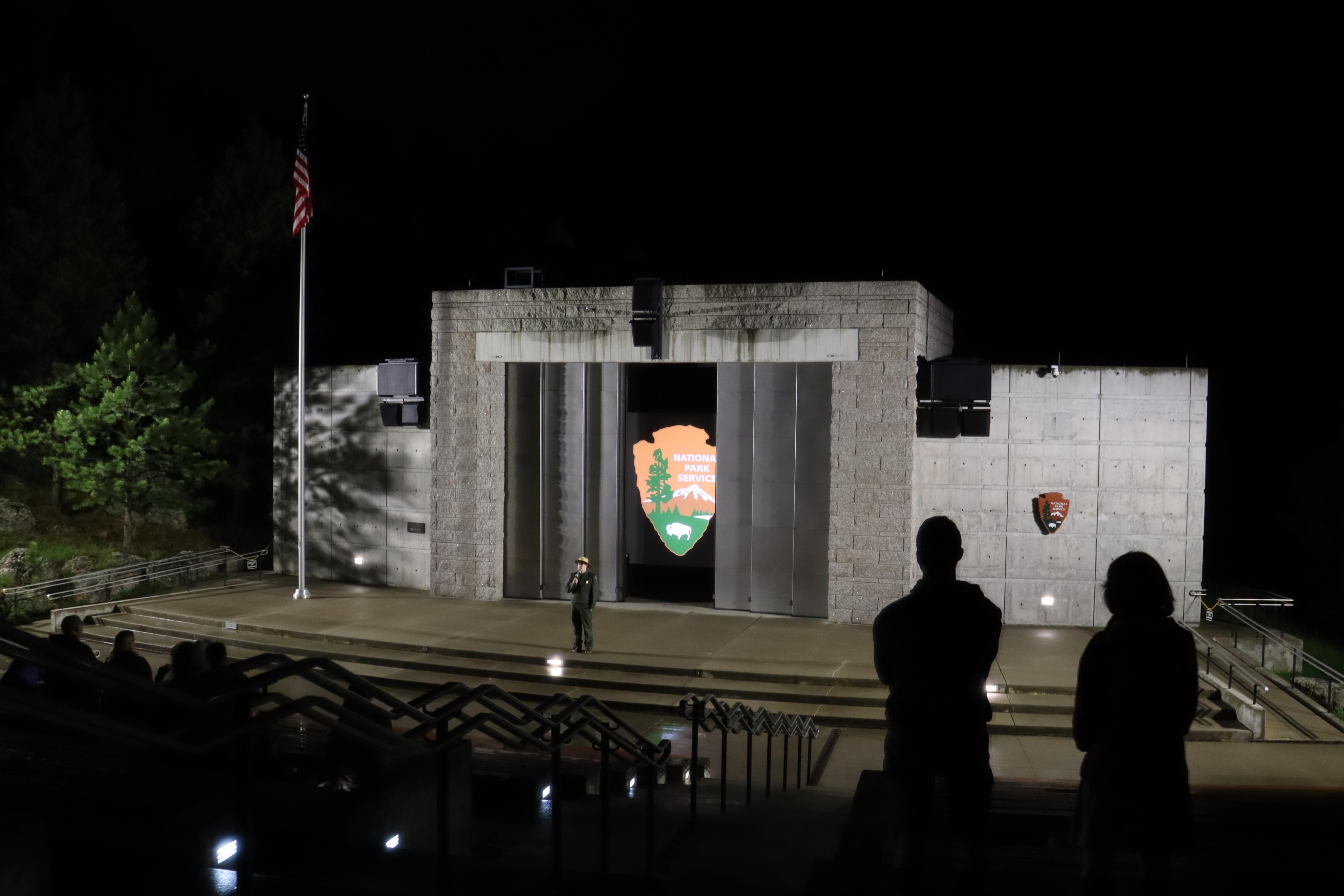 A Park Ranger stands on stage in front of a concrete building with the NPS Arrowhead on a screen.
