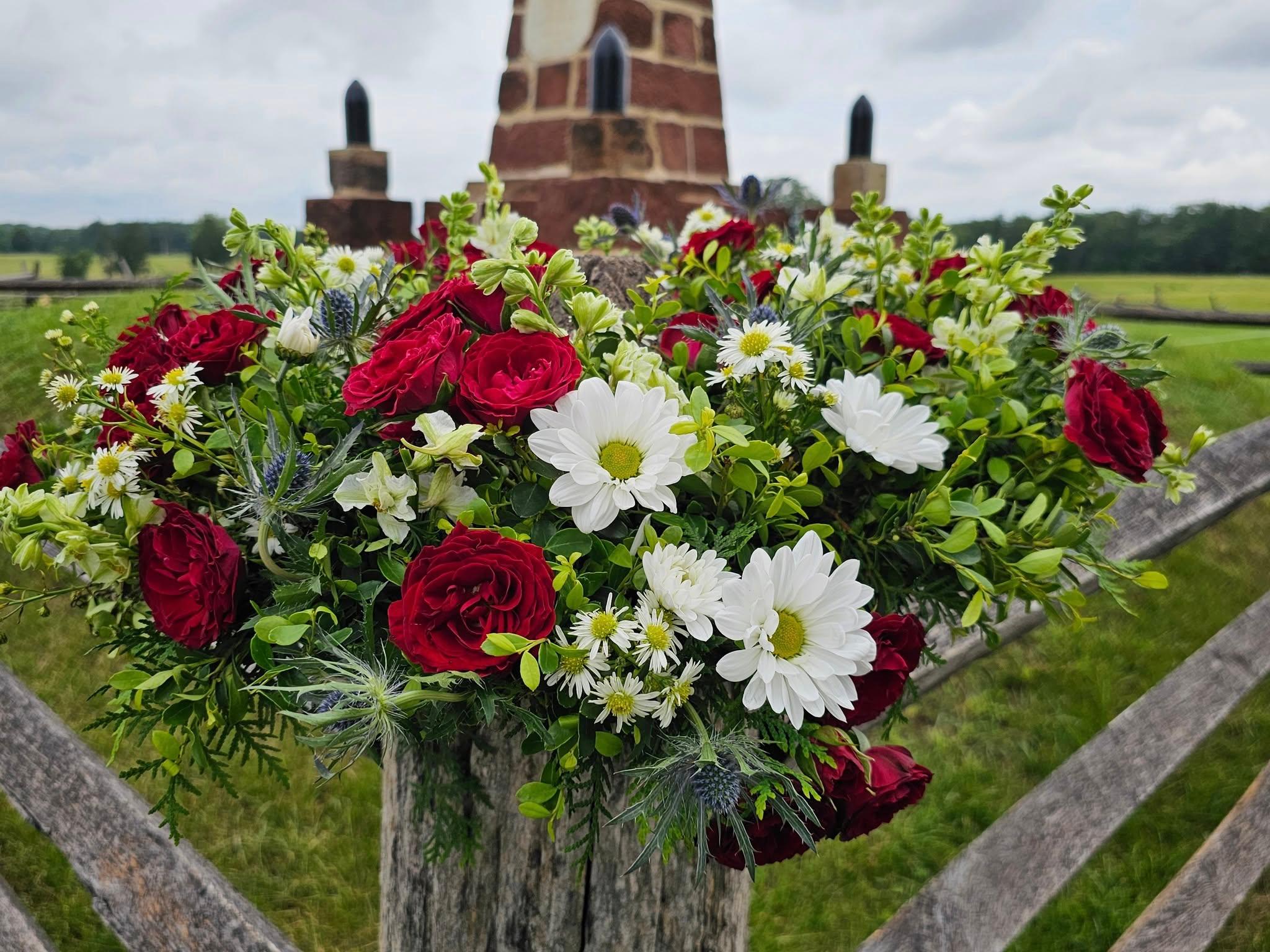 A wreath of red, white, and blue flowers sits on a fence post in front of a sandstone monument.
