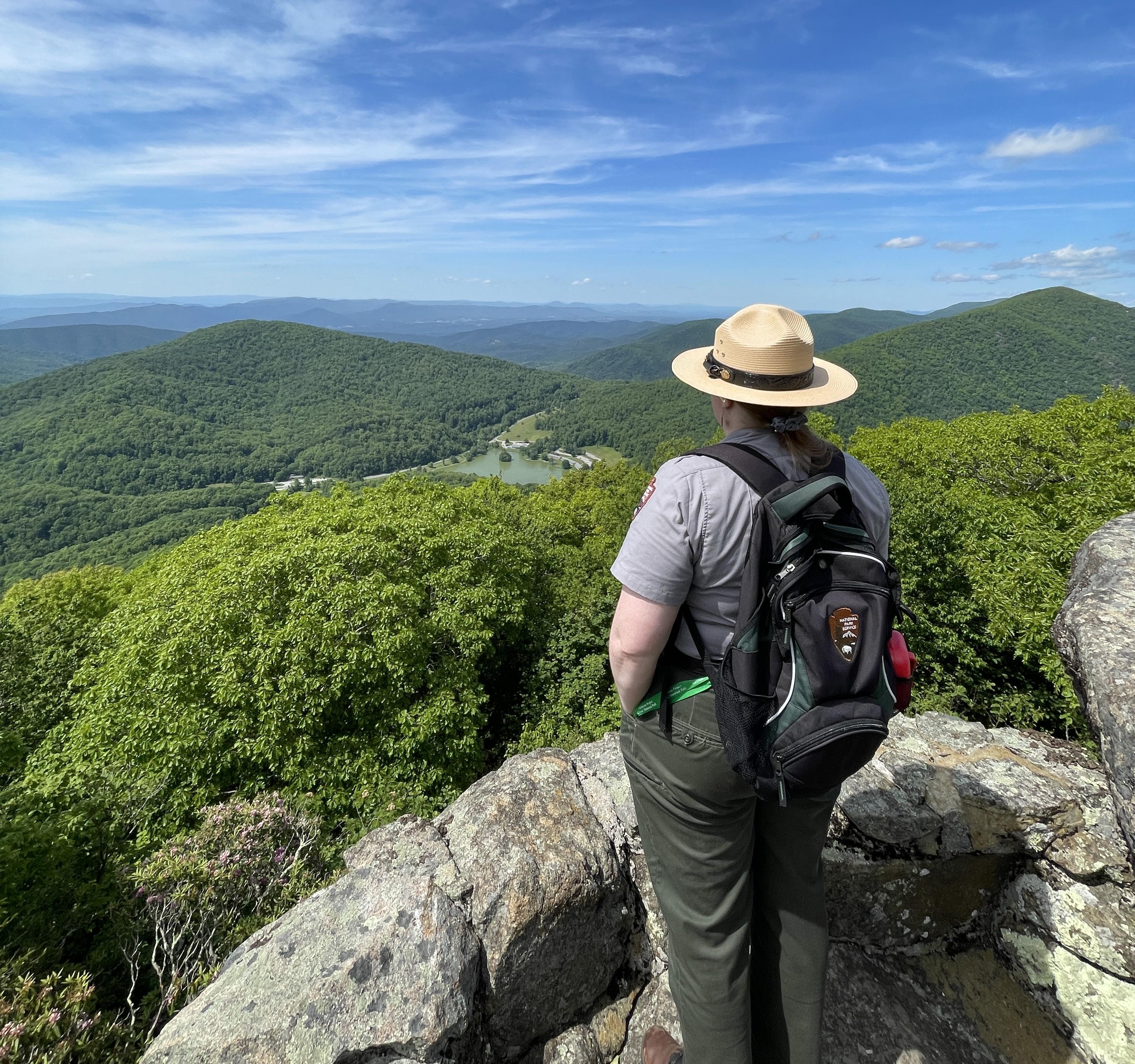 the back of a uniformed ranger looking over a lake, trees, and mountains from the top of a mountain