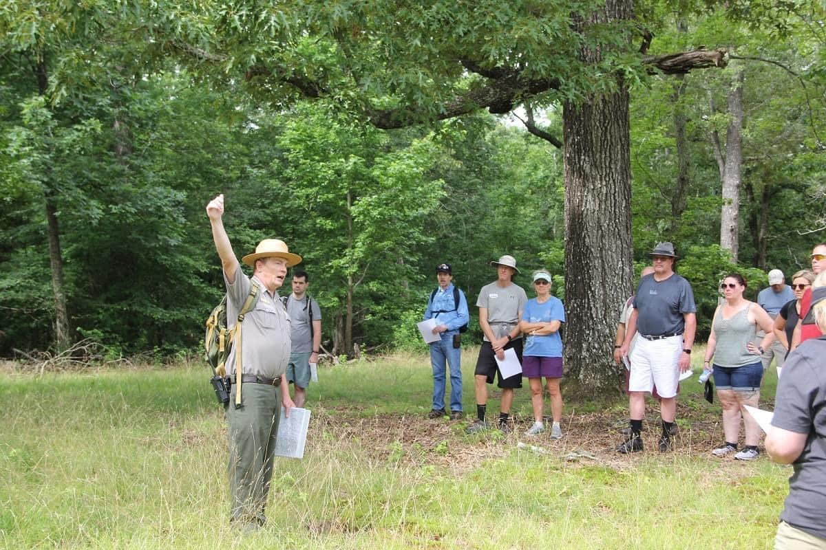 A male park ranger holds his hand above his head as he speaks to a group of visitors near a tree.