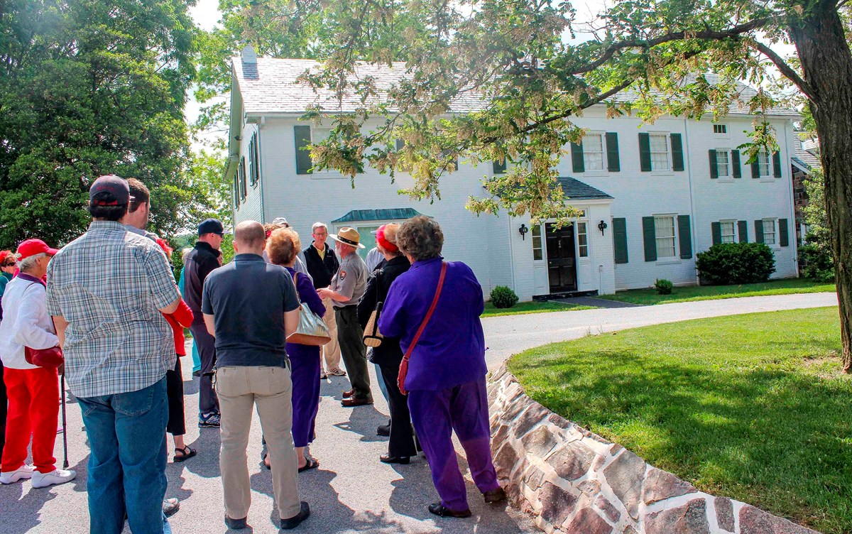 A group of visitors gather around a park ranger in front of a two-story white sided house.