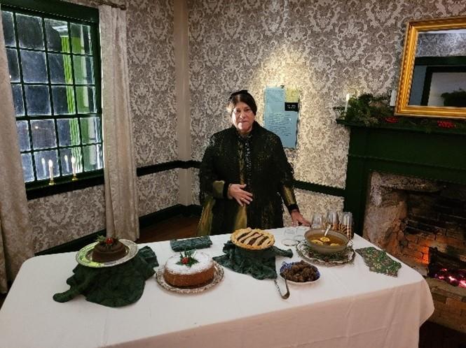 A woman in 1800s clothing stands behind a table filled with food on a white tablecloth.