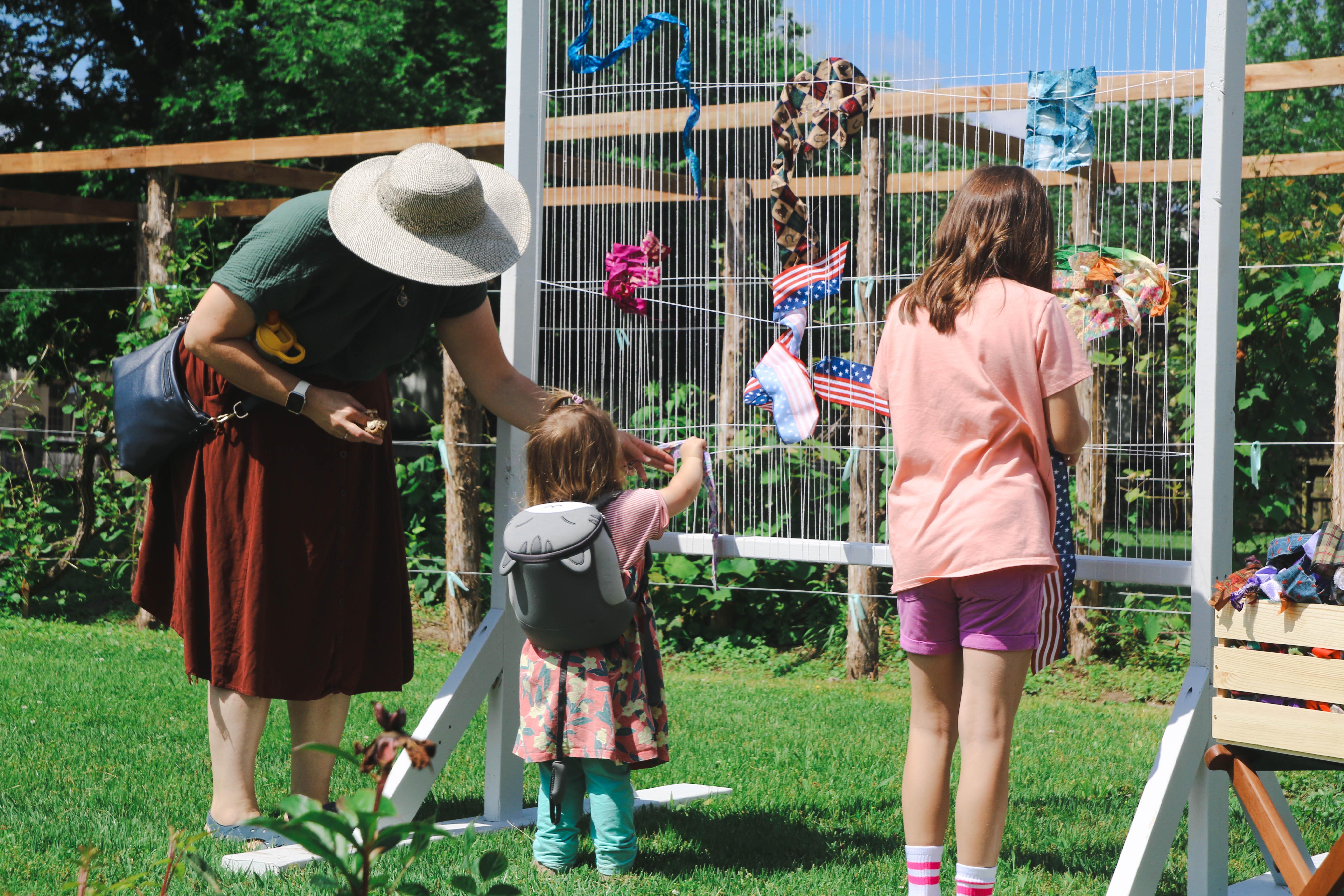 A mom and two girls stand facing a white tapestry frame tying pieces of fabric to it.