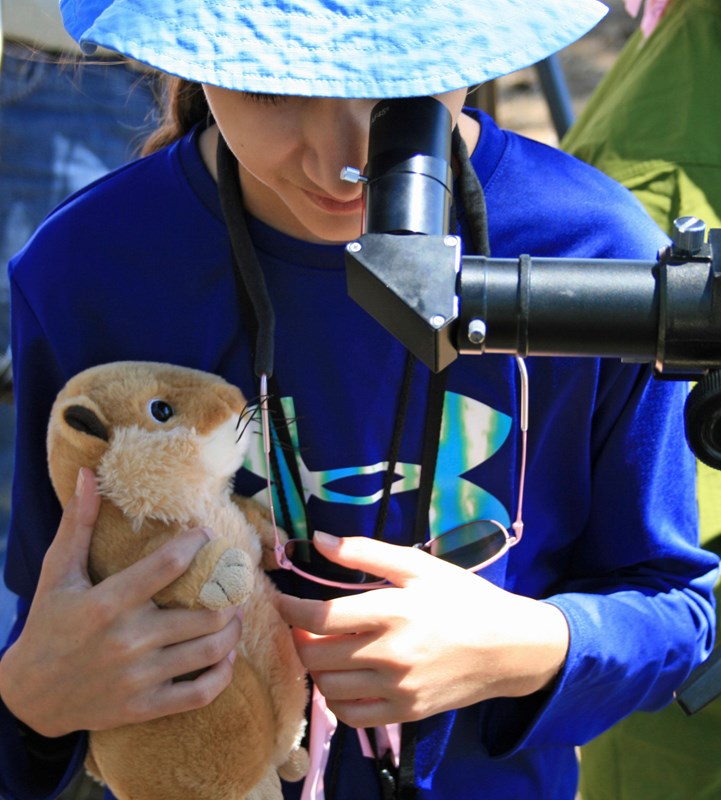 A child holding a stuffed prairie dog toy looks through a telescope eyepiece.