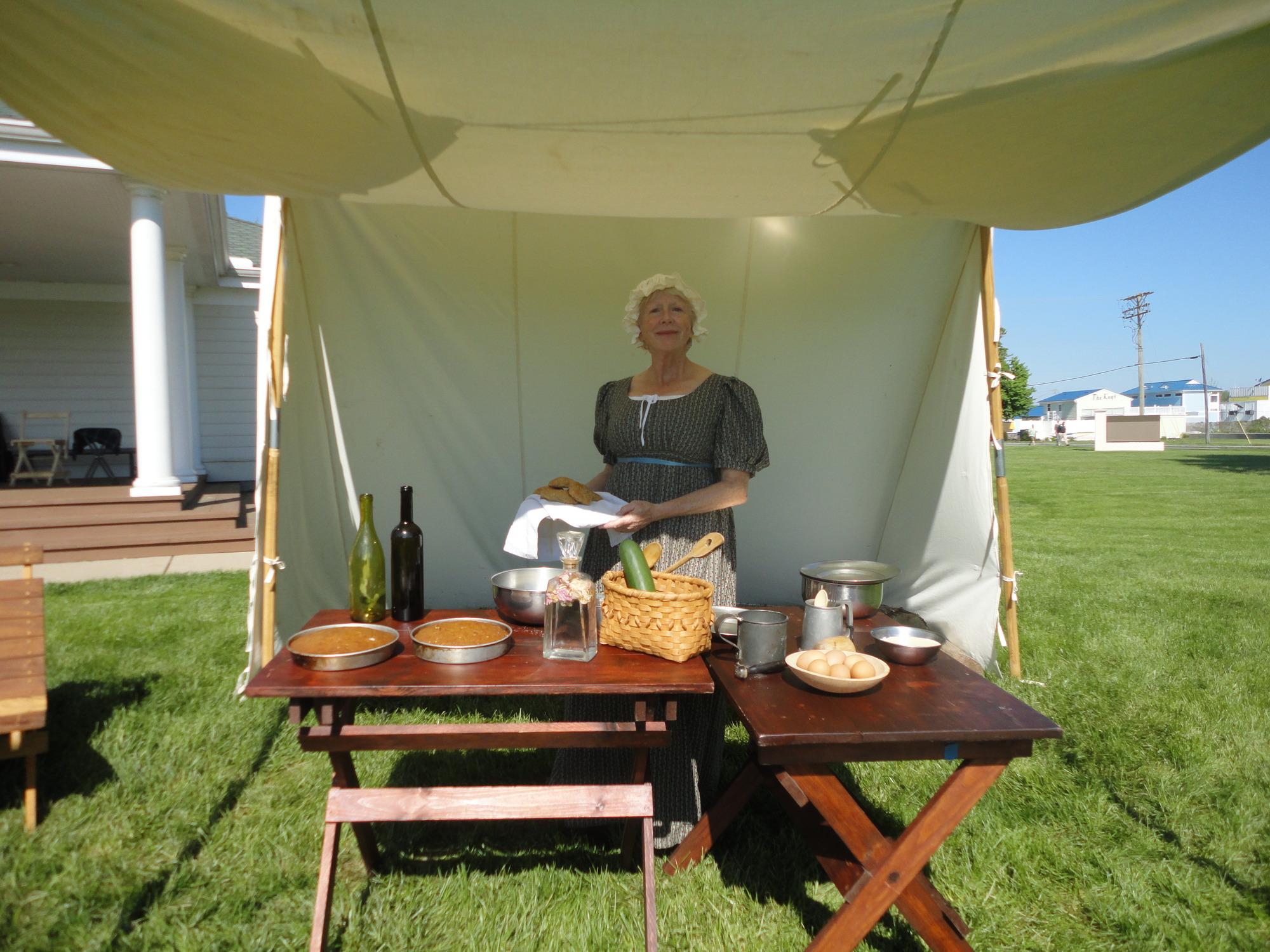 Women dressed as it is 1812 stands under a fly with food and cooking utensils.