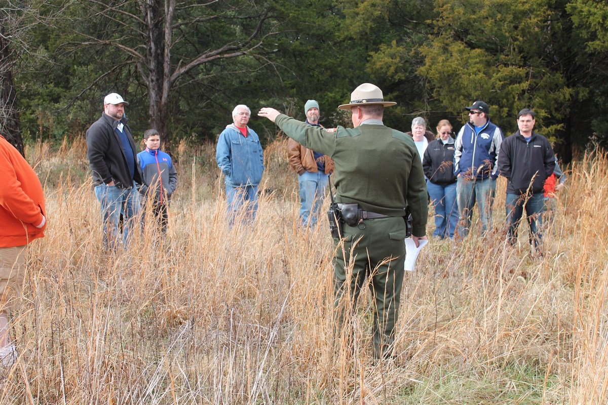 A Park ranger stands in front of people in a brown grass fields and points to the left.