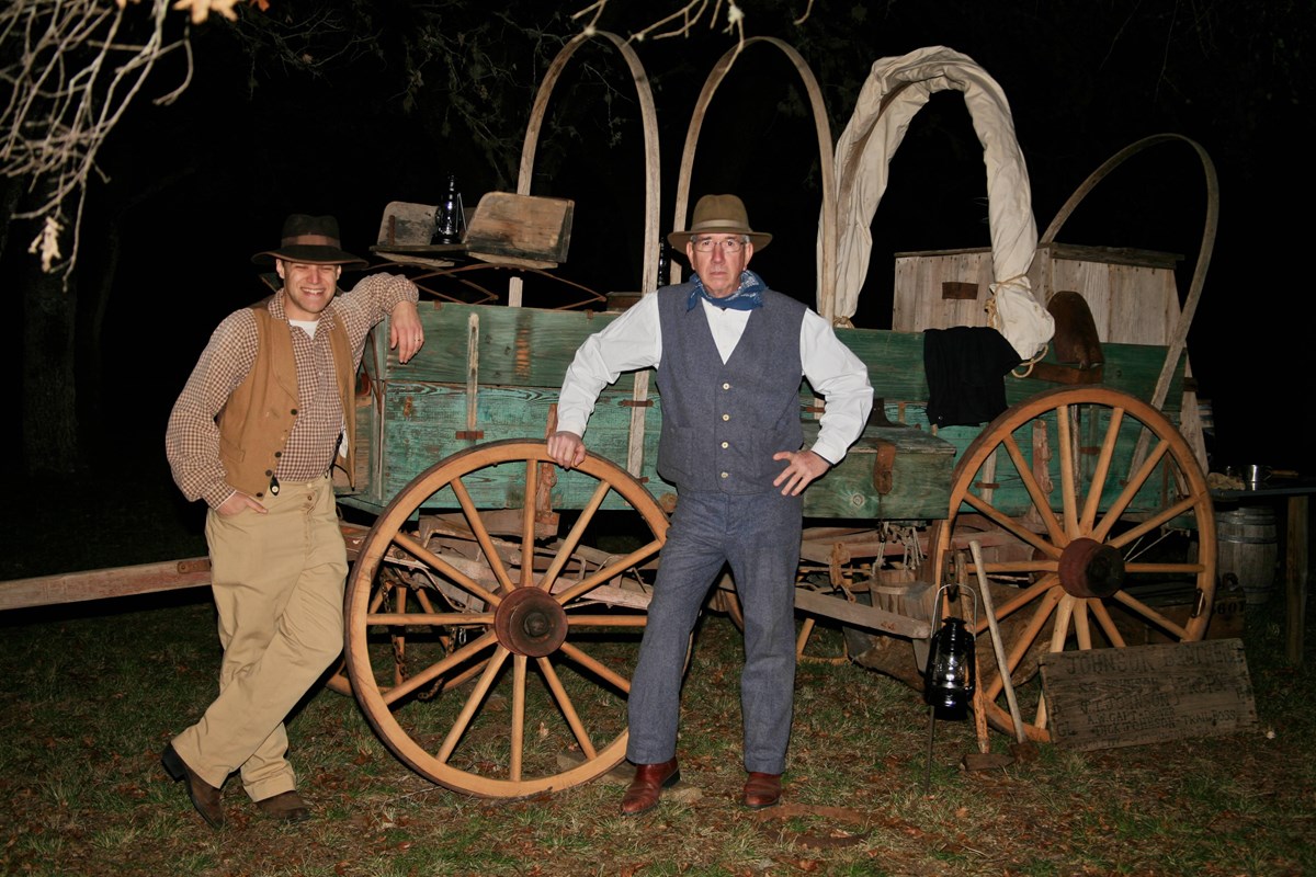 Cowboys dressed in 1860s attire stand next to a wooden chuckwagon.