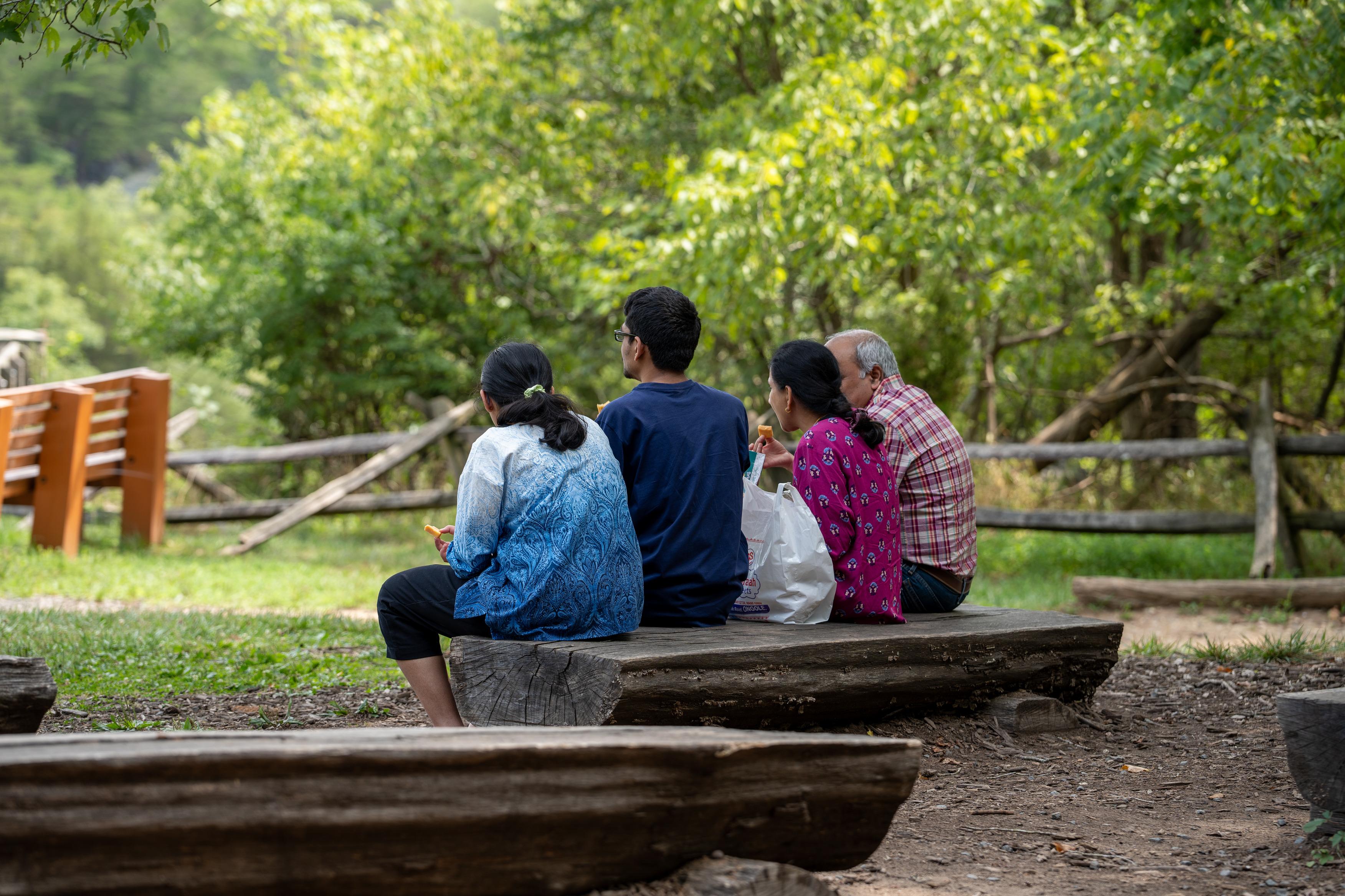 A group of four takes a break eating a snack on a bench under the shade of a tree.