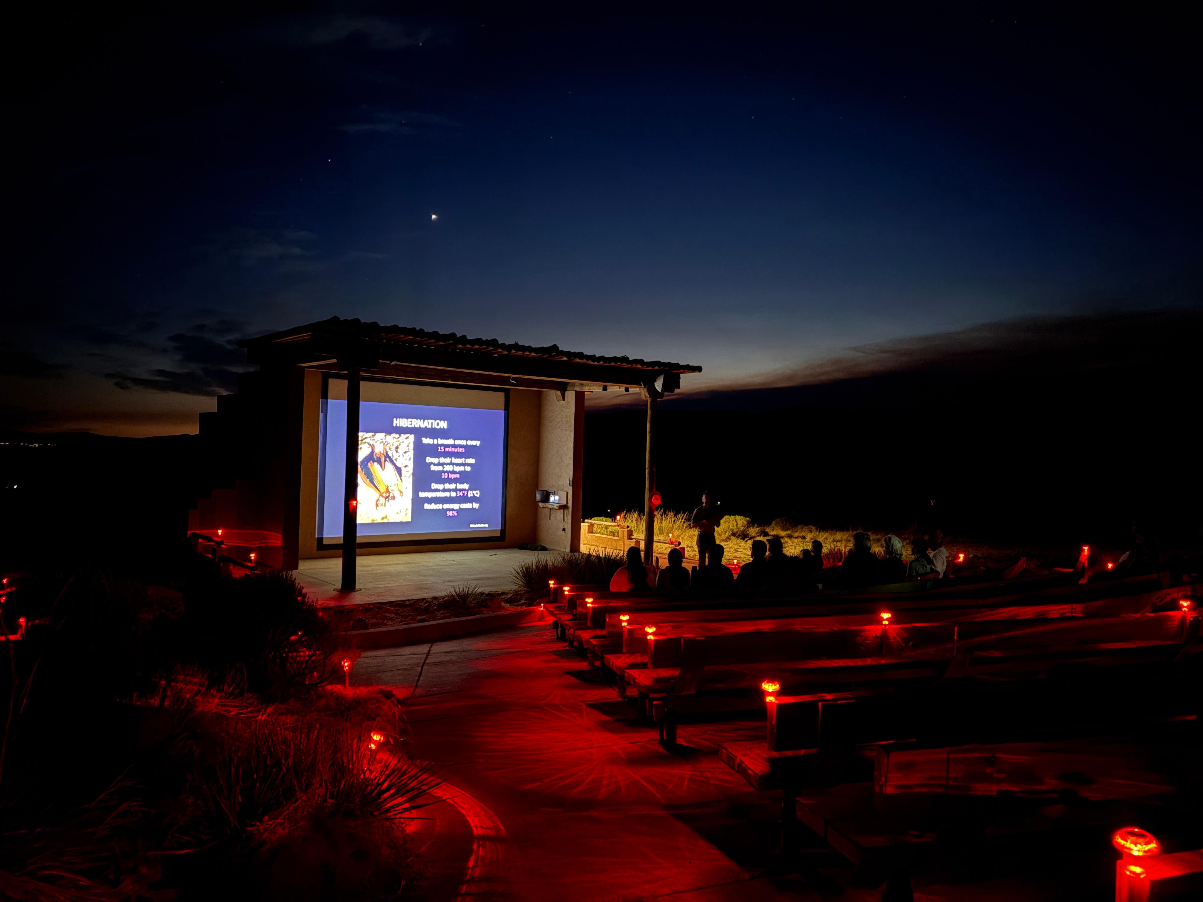 A ranger gives an evening program at an outdoor amphitheater, red lighting illuminates the seating.