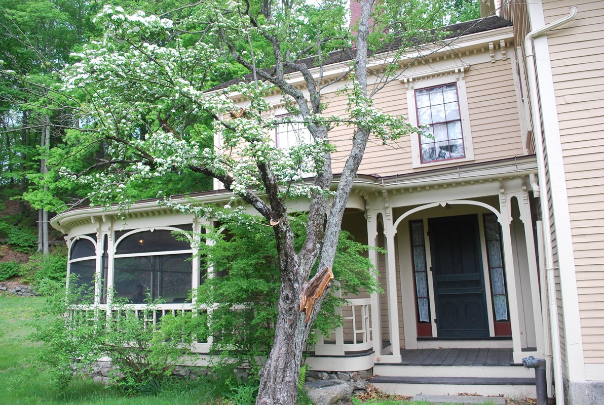 A tree-shaded porch attached to the side of a two story yellow house