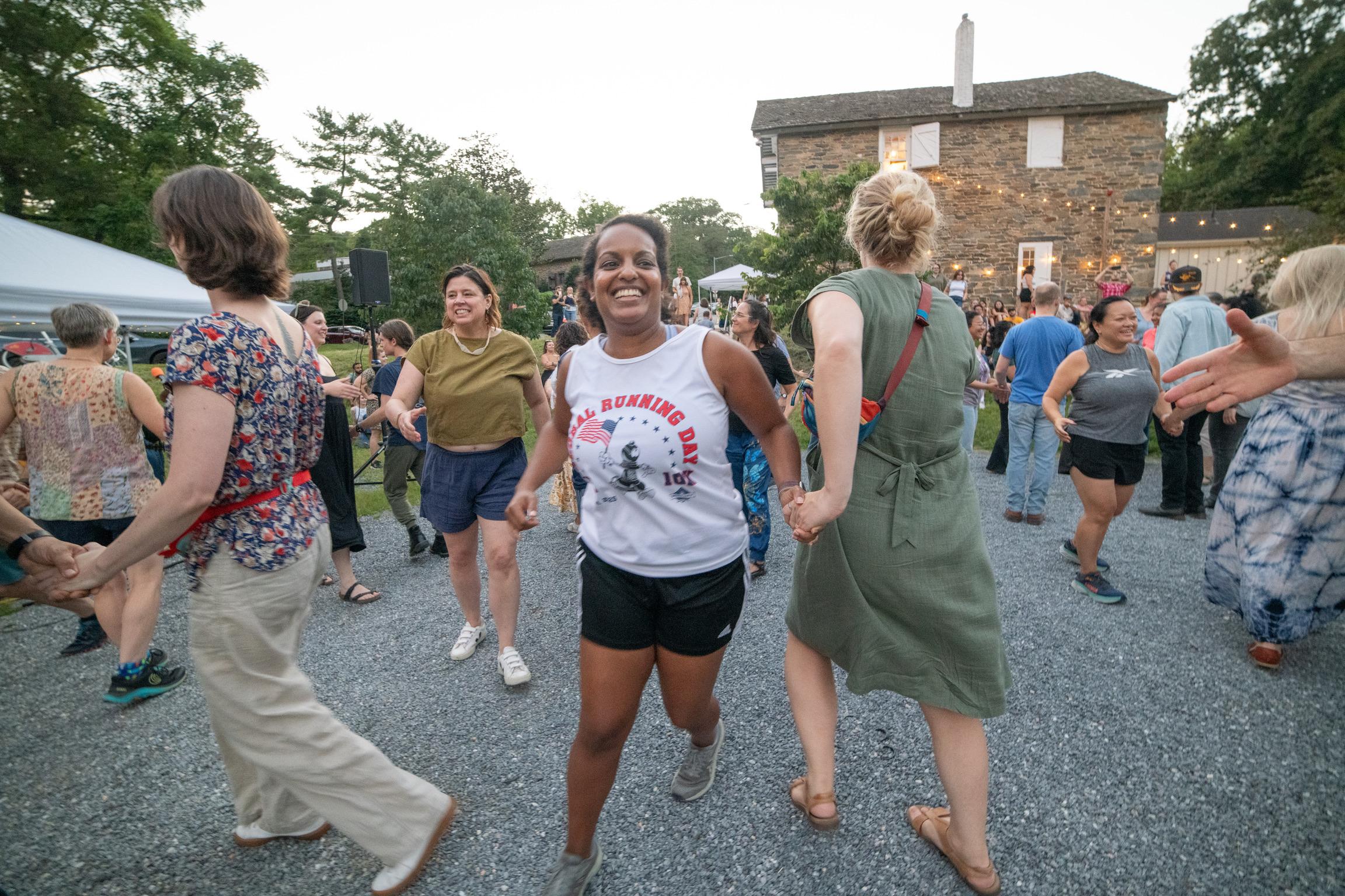 A group of people are dancing on gravel in front of a historic stone barn.