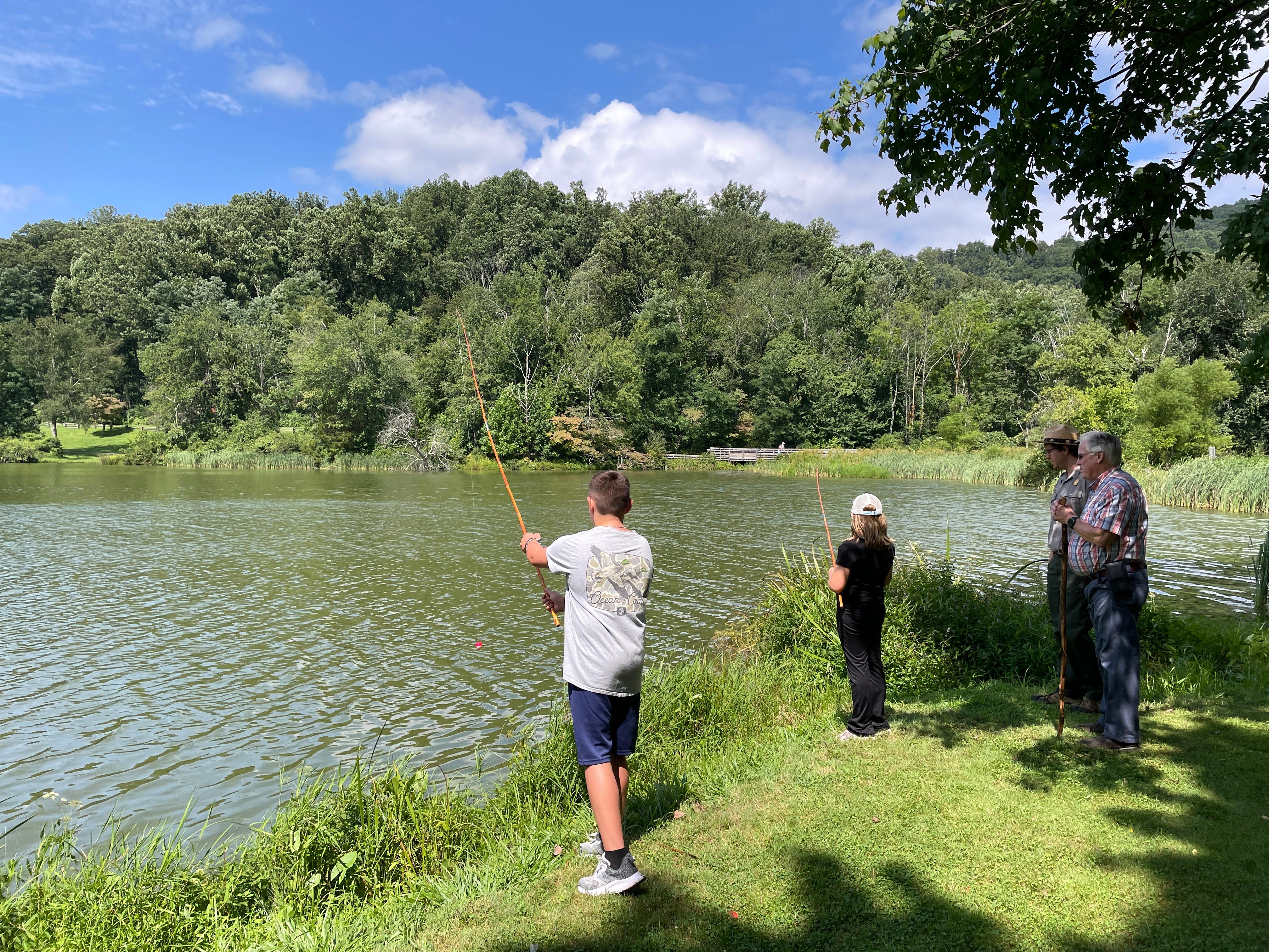 2 kids fishing along the shoreline of a pond surrounded by trees and greenery